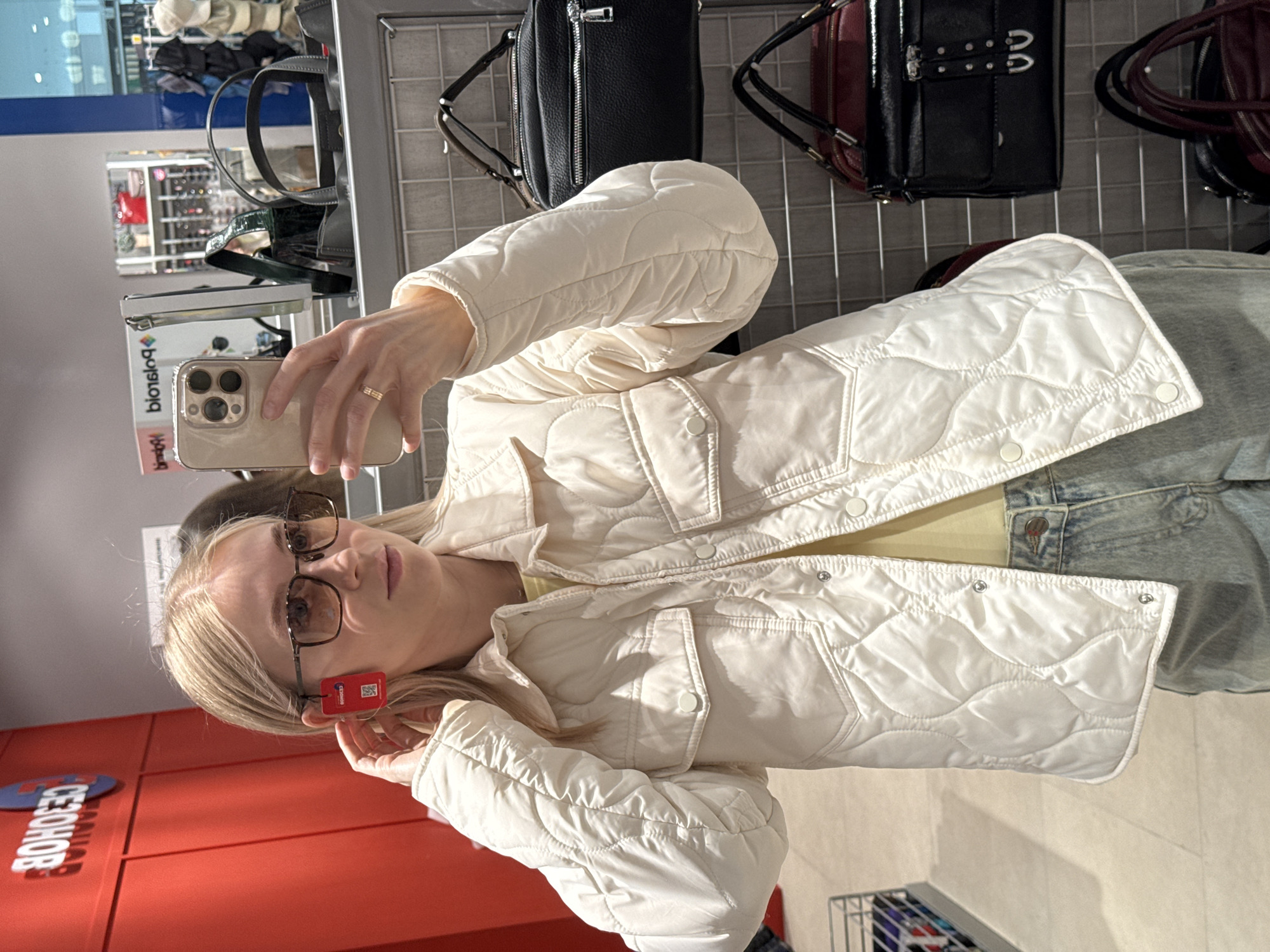 A woman with blond hair and glasses is taking a selfie in a store, presumably in Izhevsk, Russia, based on the “CEOHOB” (чехолов) signage. She is holding a phone in her left hand and facing the camera. The background consists of retail shelving with various handbags on display. The woman is wearing a light-colored quilted jacket. The environment appears to be a retail setting.