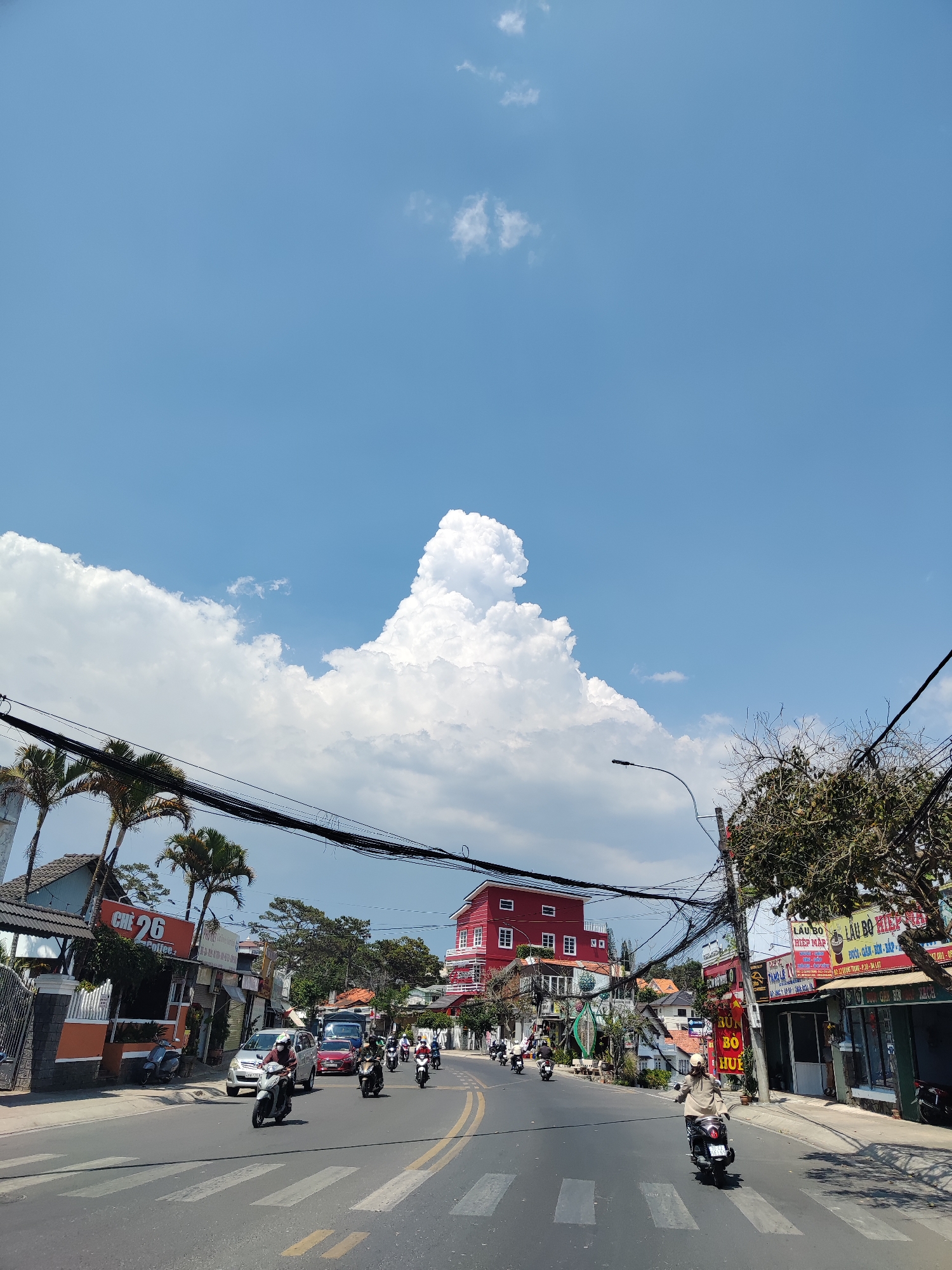 The image shows a street scene in Da Lat, Vietnam, under a bright blue sky with large cumulus clouds. A main road is visible, with two lanes and zebra crossings. Motorbikes are the predominant form of transportation. Various buildings line the street, including a restaurant labeled "CHE 26 Coffee", a bright red building further up the road, and "LAU BO HIỆP MAP LAU BO DEP" restaurant. Power lines crisscross the sky above the street. Palm trees and other greenery are interspersed among the buildings.