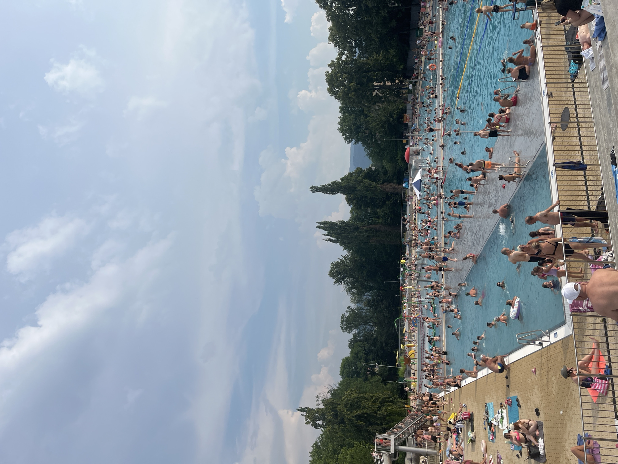 The image shows a crowded outdoor swimming pool on a sunny day. The pool is filled with people of all ages swimming, floating on inflatable rings, and relaxing on the pool deck. The water is a bright blue, and the pool deck is paved with yellow tiles. Surrounding the pool are lush green trees, and in the background, there are rolling hills. The sky is a clear blue with a few white clouds. The atmosphere appears to be festive and relaxed, with many people enjoying the warm weather and recreational activities. It is difficult to determine the exact time of day, but the lighting suggests it is daytime, likely mid-morning or afternoon. There are no visible text elements in the image.