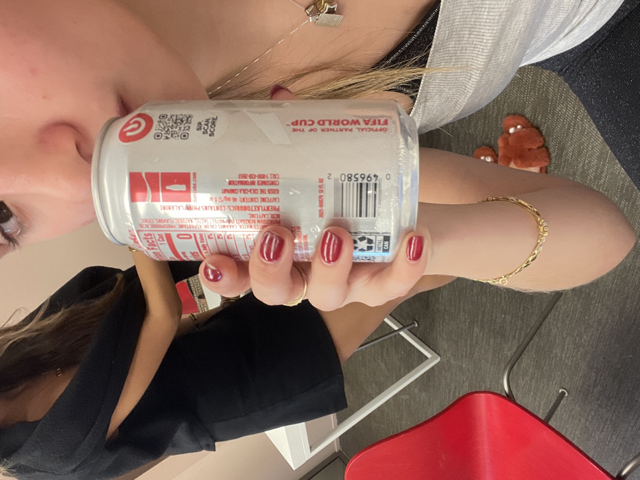 A person holds a Diet Coke can featuring information like "OFFICIAL PARTNER OF THE FIFA WORLD CUP." The can displays nutrition facts, ingredients like aspartame and caffeine, and consumer information including a phone number and website. The person has dark red nail polish. A gold bracelet is on the wrist. Red shoes and a white table are visible in the background.