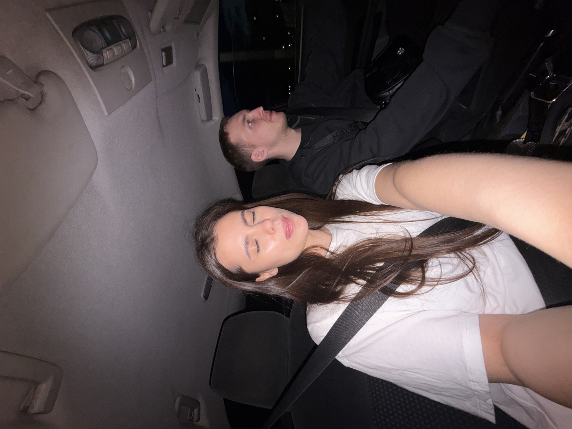 Two individuals are seated inside a vehicle at night, presumably in Chaikovsky, Russia, as indicated by the prompt's context. The person in the foreground, a young woman with long dark hair, is wearing a white t-shirt and is looking towards the camera with her eyes closed and tongue slightly extended. A black seatbelt is visible across her chest. To her right, a young man with short brown hair is looking out the side window. He is wearing a black hooded jacket and has a black bag slung across his chest. The interior of the car, including the ceiling and seats, is visible. The background outside the vehicle is dark, with faint indications of lights suggesting an urban or suburban environment.