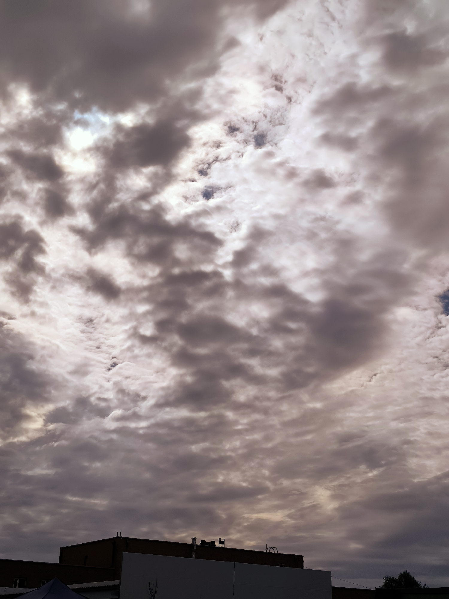 The image shows a sky filled with dramatic, textured clouds. Some clouds are dark and ominous, while others are lighter and allow patches of sunlight to break through. The clouds create a sense of depth and movement in the sky. Below the clouds, the tops of several buildings are visible, suggesting an urban environment. The architecture appears to be functional rather than ornate. The overall impression is of a cloudy day, possibly late afternoon or early evening, with the light diffused by the cloud cover. There is no direct indication of the specific location within Karaganda, Kazakhstan.