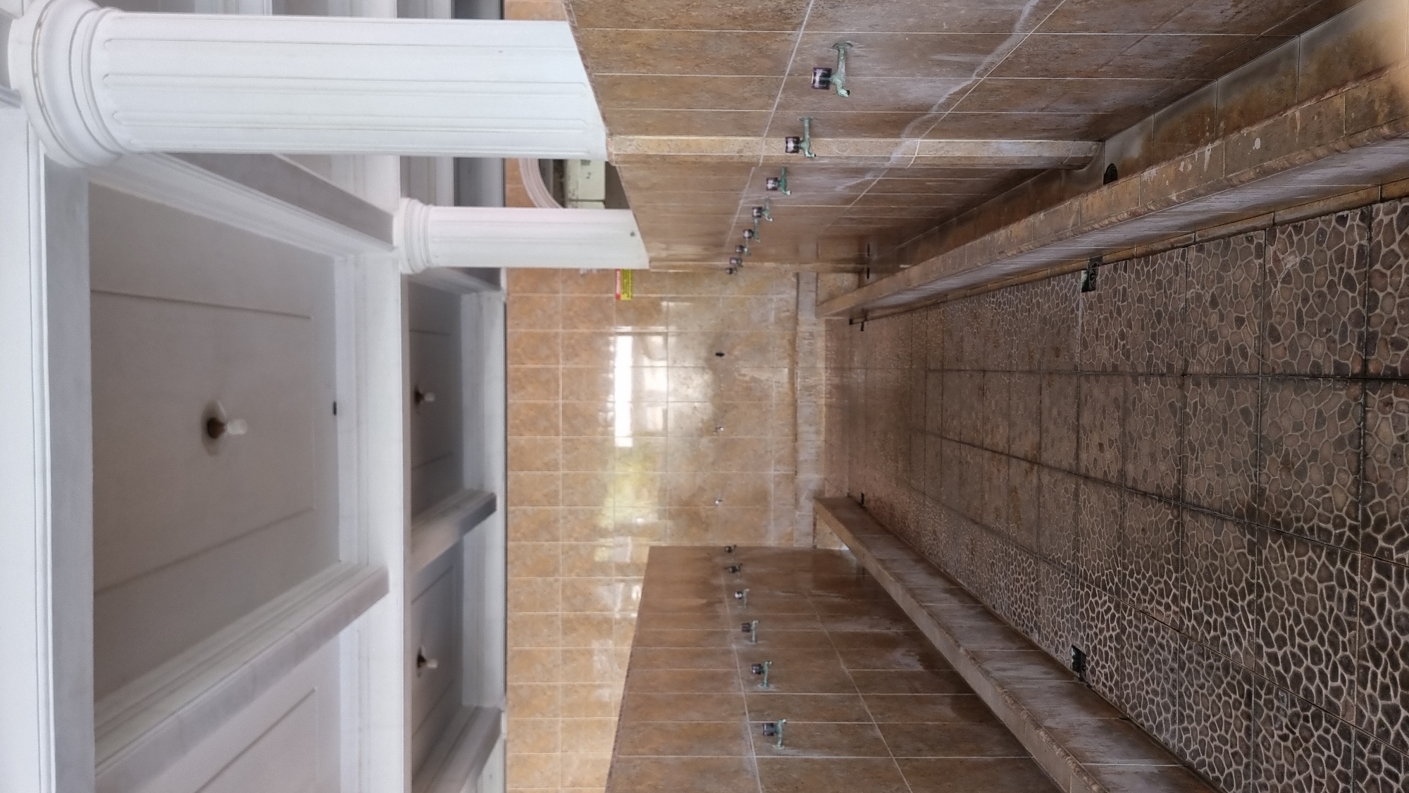 This image shows a communal ablution area, likely at a mosque or similar religious site, in Keudekruenggeukueh, Indonesia. The space is open-air, indicated by the natural light illuminating the tiled walls and wet floor. On either side of a central pathway, low concrete basins are lined with faucets for washing. The walls are covered in light brown tiles, and the floor is a mosaic of darker, irregularly shaped tiles, some of which appear wet. The ceiling is ornate, with white beams and recessed panels, and a few bare lightbulbs are visible. Large white fluted columns support the ceiling structure. No people are visible in the image, and there are no discernable actions or interactions taking place. The scene is static and focuses on the functional design of the ablution facility.