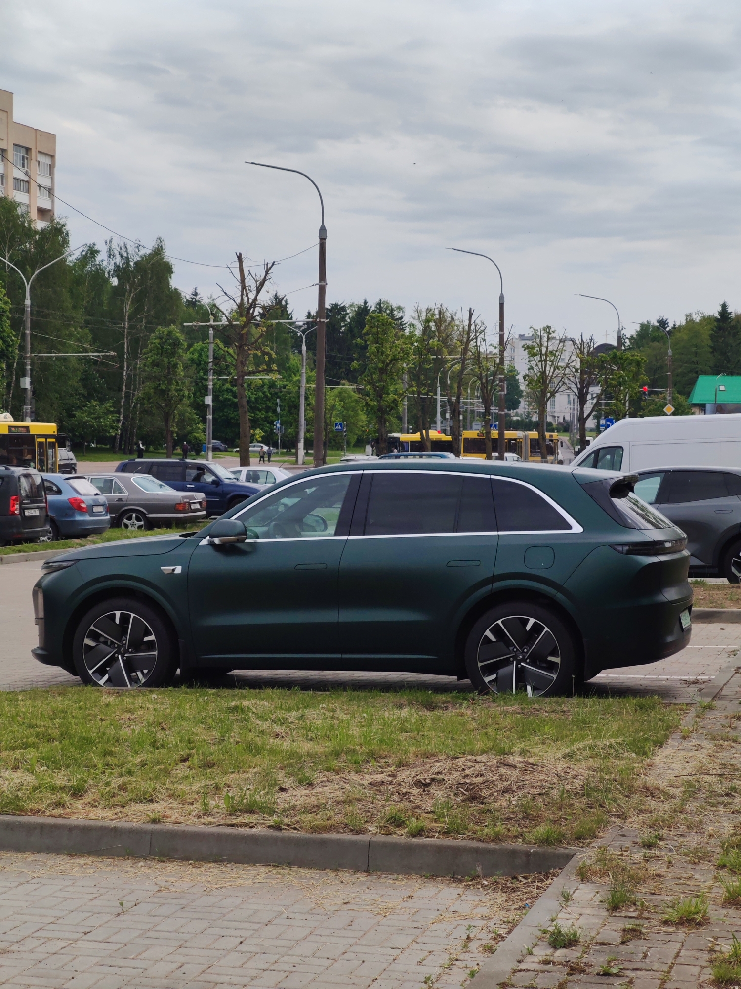 The image shows a dark green SUV parked on the edge of a paved area next to a grassy median. The location appears to be an urban area, as suggested by the nearby buildings, streetlights, and other parked vehicles. The weather seems overcast. Specific details of the city or any recognizable people are not evident in the image.