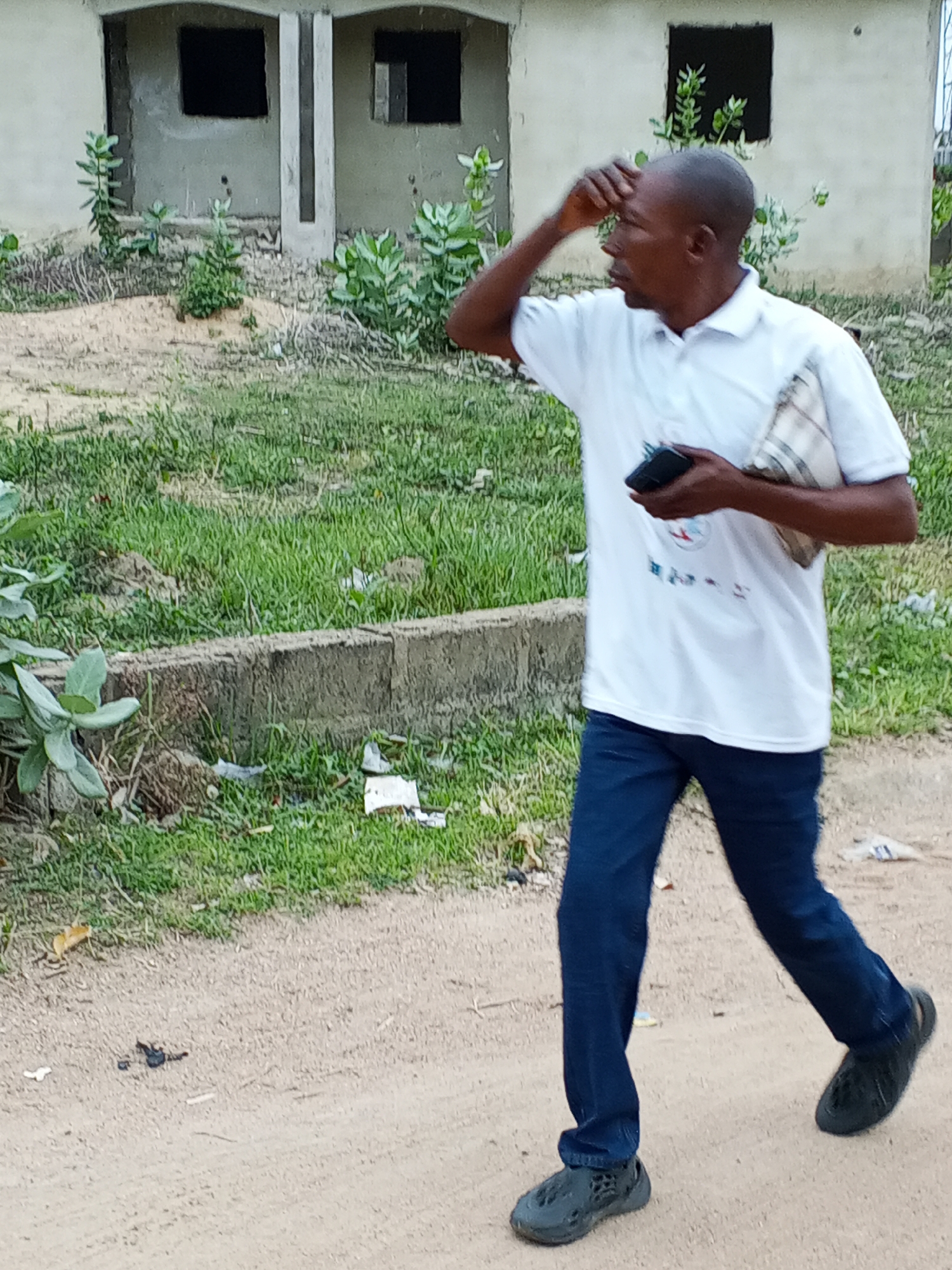 A man is walking along a dirt path in Otokiti, Nigeria. He is dark-skinned, balding, wearing a white polo shirt, dark blue jeans, and black shoes. He holds a dark-colored cell phone in his left hand and has a light-colored, patterned cloth slung over his left shoulder. His right hand is raised to his forehead, as if shielding his eyes. To his left is green grass and a low concrete wall with scattered litter. Behind him and to the left is a rough, unfinished building with open window spaces. The ground is sandy and there is trash scattered about. The location is Otokiti, Nigeria.