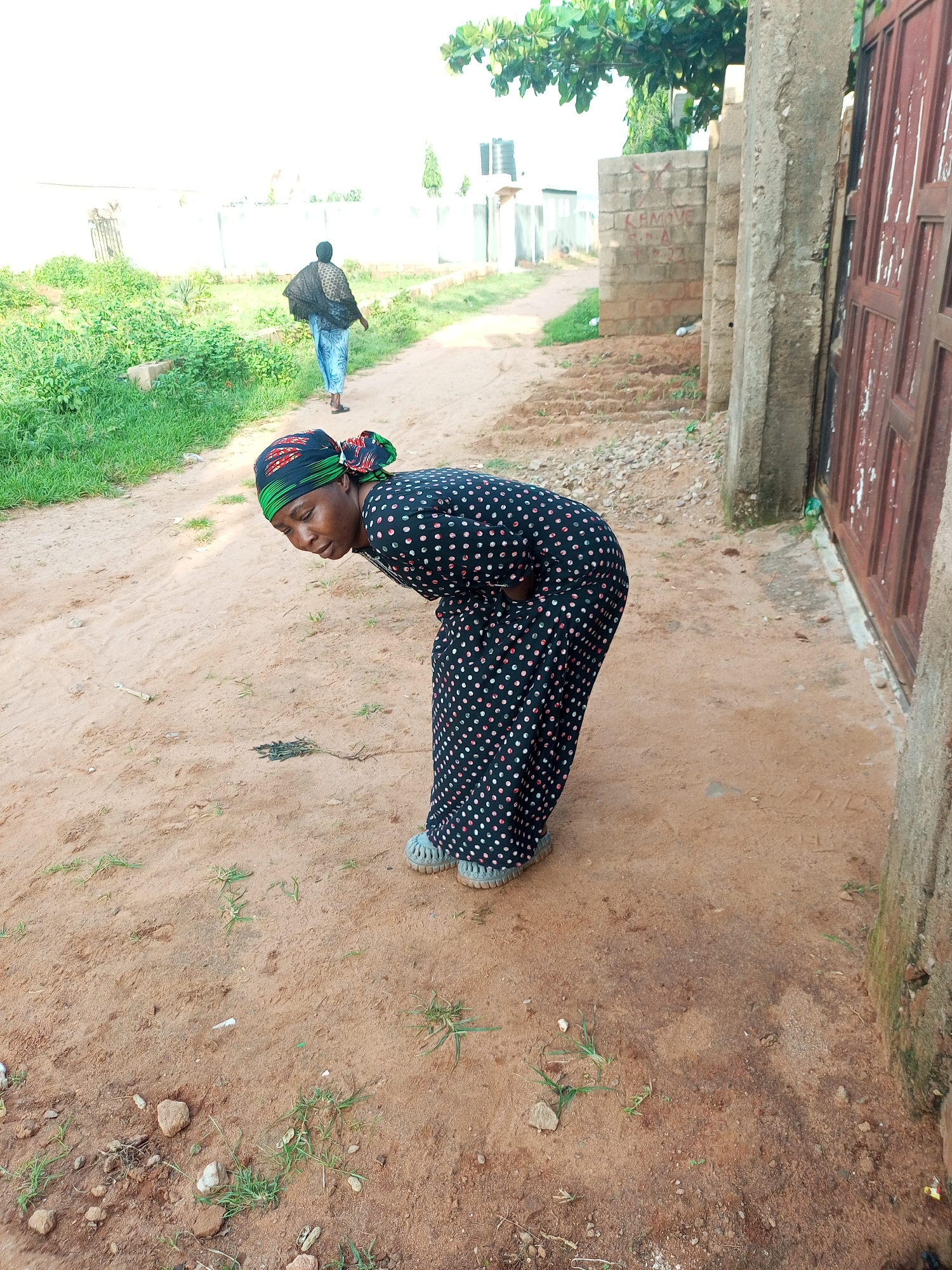 In the city of Tempure, Nigeria, a woman is bending over on a dirt path. She is wearing a black dress with white polka dots and a headscarf with green and black patterns. Her head is bowed, and she appears to be looking at something on the ground. In the background, another person is walking away from the camera. The environment is dusty with patches of grass and some debris. A concrete wall and a rusty metal gate are visible to the right of the frame.