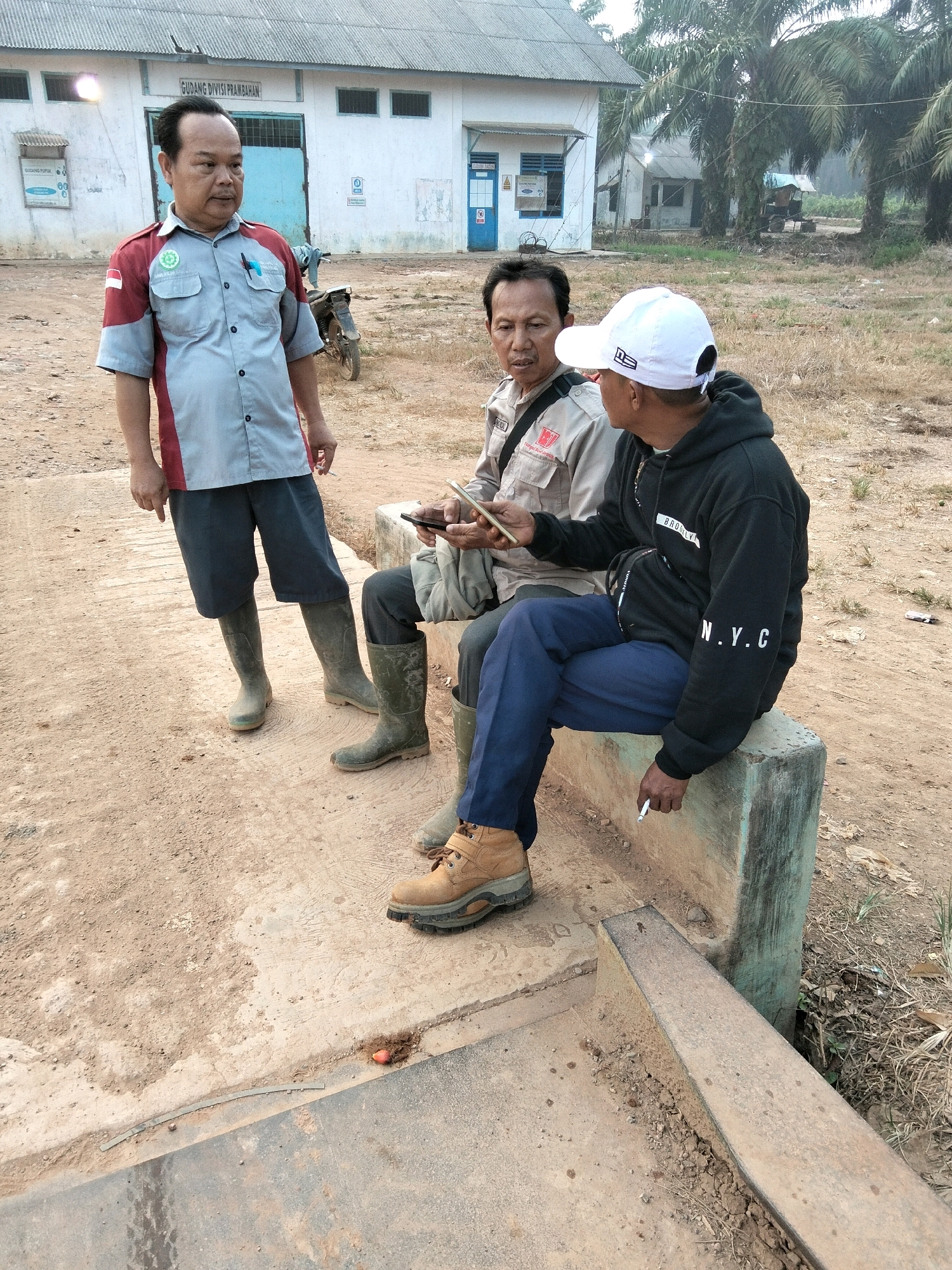 The image depicts an outdoor scene at what appears to be a warehouse or industrial facility in Teluk Tenggirik, Indonesia. The central focus is a group of three men in conversation. One man, standing and wearing a grey and red collared shirt, dark shorts, and green rubber boots, is gesturing while speaking. To his right, two men are seated on a concrete structure. One seated man, wearing a light-colored collared shirt and dark pants, holds a mobile phone. The other seated man wears a black hoodie with "BROOKLYN" and "N.Y.C." visible on the sleeves, blue pants, and tan work boots. He is holding a cigarette. The background features a white building with the text "GUDANG DIVISI PRAMBAHAN" visible on it, along with palm trees and dry, dusty ground. A motorcycle is parked behind the standing man. The overall environment is rural and appears to be a work site.