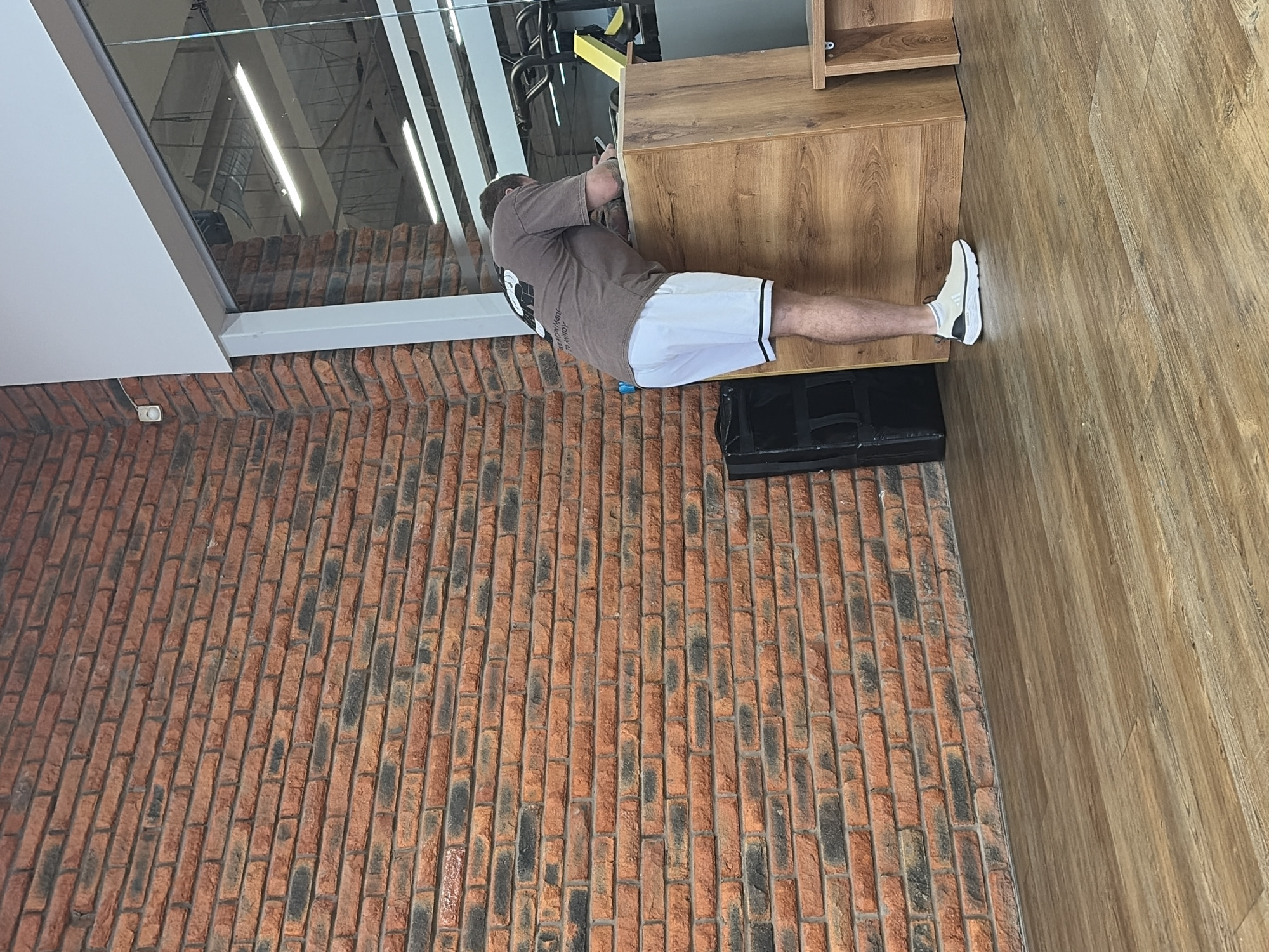 A vertically oriented photograph depicts a man indoors performing an exercise involving a wooden cabinet-like structure. The man wears a brown t-shirt and white athletic shorts with black trim. He is leaning onto the cabinet with his hands, while his legs extend away from the cabinet, with his right foot touching the wooden floor. A black rectangular object rests near his legs on the brick floor. The floor is made of red bricks in the bottom left portion and is finished with a light brown wood pattern on the right. A window reflecting overhead lighting and gym equipment is visible above and to the left of the man. Based on the mention of the city of Chaikovsky, Russia, the scene is likely located in a gym or fitness facility within that city.