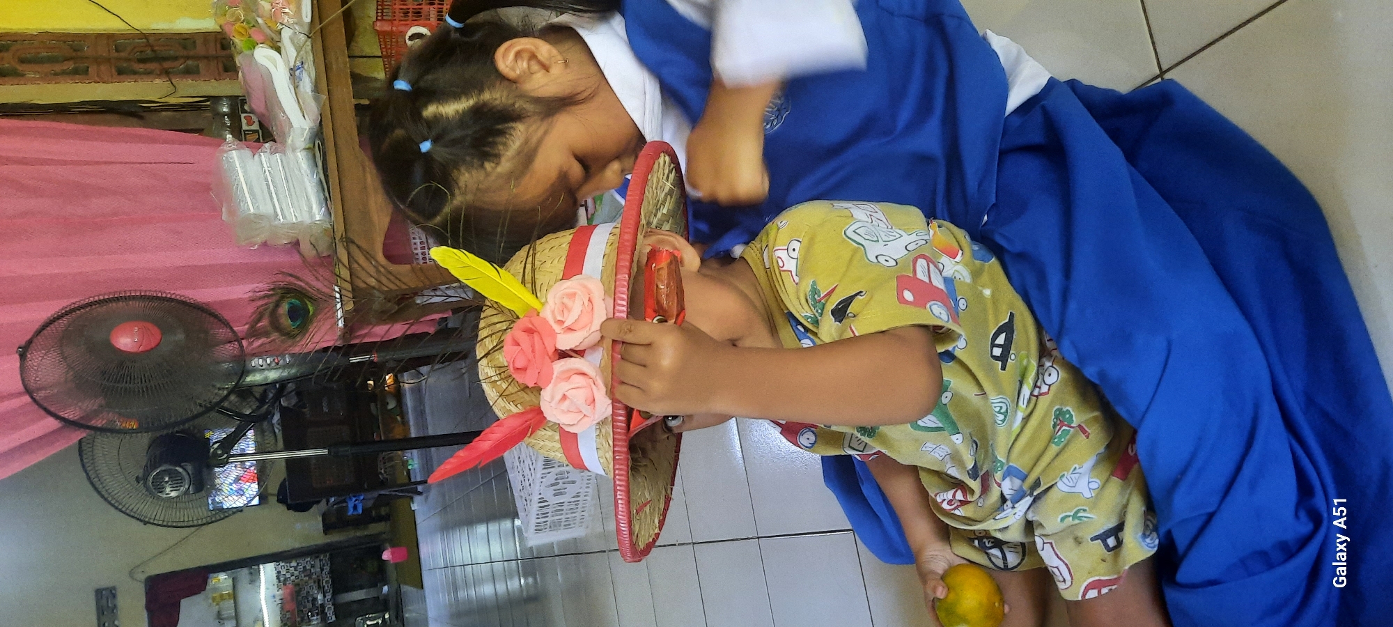 In a brightly lit indoor setting, possibly a home or school room in Ploso, Indonesia, two young children are interacting. A boy, wearing a yellow t-shirt with a car print and a straw hat adorned with pink fabric flowers and colorful feathers, sits with an orange in his left hand. He is looking through what appear to be red toy glasses. He is being held by an older girl, who is wearing a blue and white uniform. Her head is bowed, and her right arm is reaching towards the boy's hat. The background includes a large black oscillating fan, a pink curtain, and a table with stacked white items. The flooring is tiled. The Samsung Galaxy A51 watermark is visible in the bottom left corner.