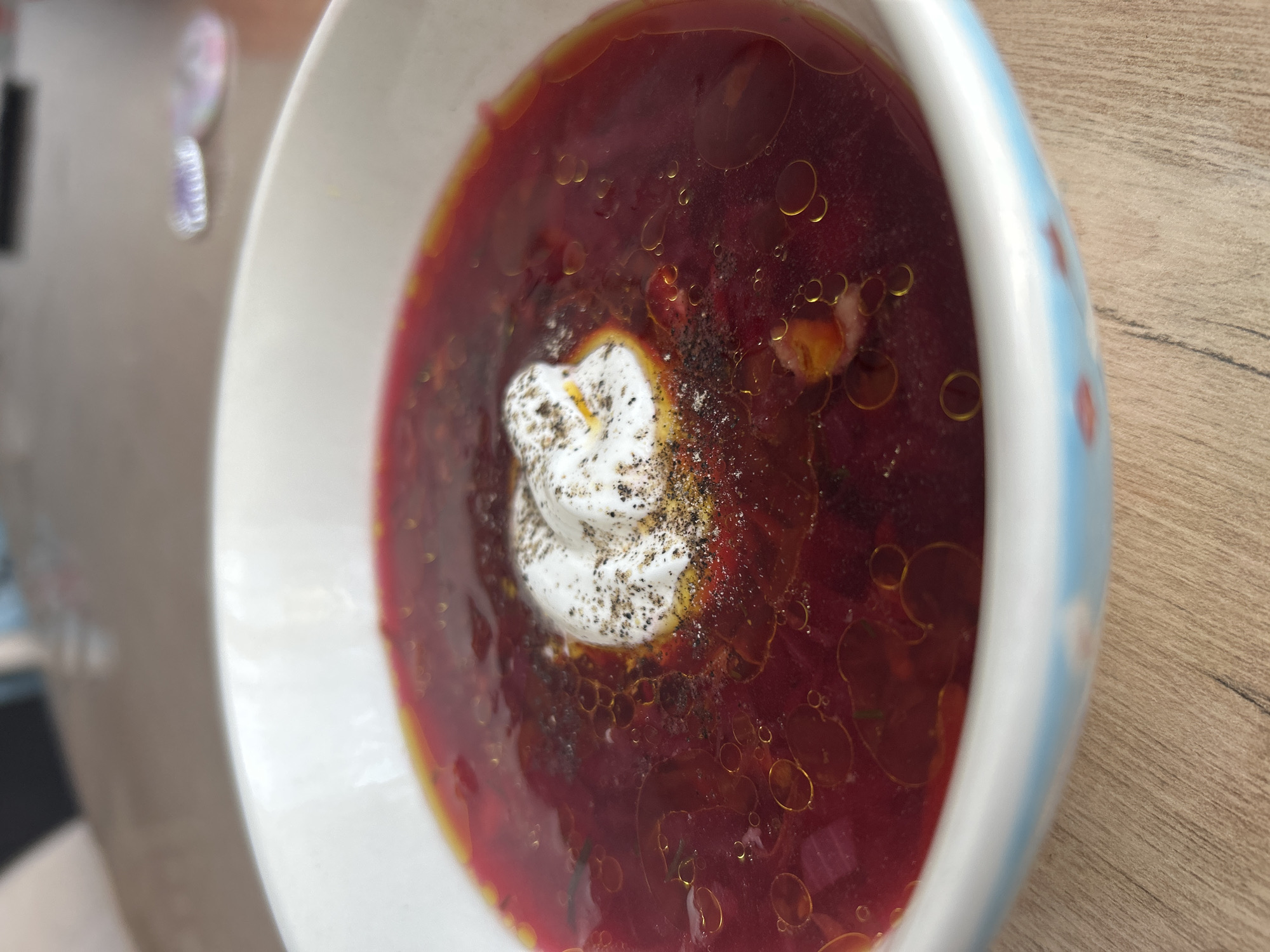 The image shows a bowl of borscht, a beet soup, topped with a dollop of sour cream and sprinkled with pepper. The soup is reddish-purple in color and has small droplets of oil on the surface. The bowl is white with a light blue accent band along the outer edge. The bowl sits on a wooden surface. There are no people visible in the image. The location is not discernible from the image itself. Therefore, I cannot provide information about the location being Chaikovsky, Russia.