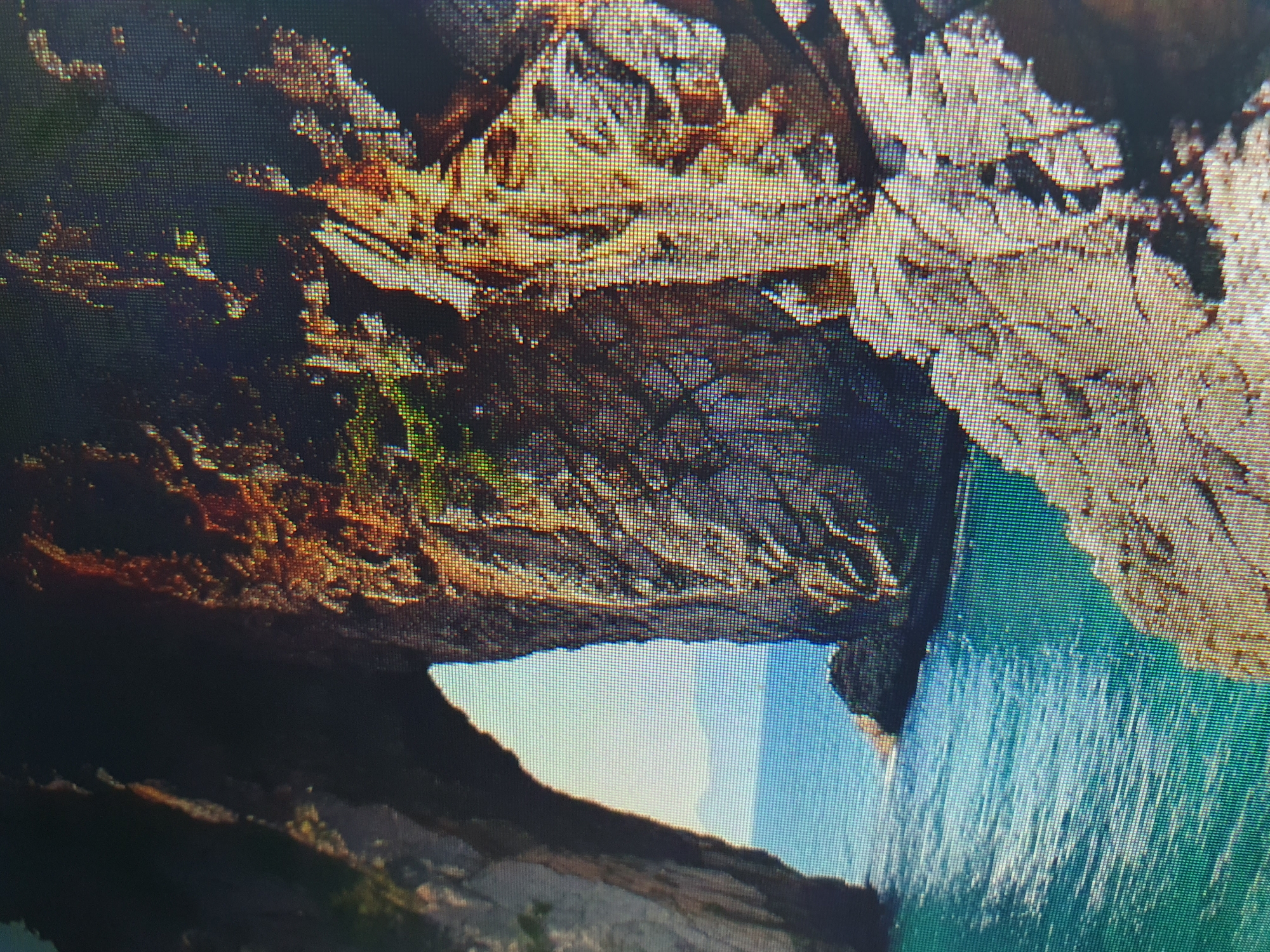 The image shows a rocky coastal landscape with a natural archway. The water is a vibrant turquoise, and the rocks are a mix of dark brown and lighter tan colors, with some vegetation growing on them. The sky is a pale blue, with faint mountains visible in the distance through the arch. The image appears to be a photograph, likely taken during the daytime, with the sun illuminating the scene. There are no people or man-made structures visible in the image. The location is not in Tashkent, Uzbekistan, as it is a coastal scene.