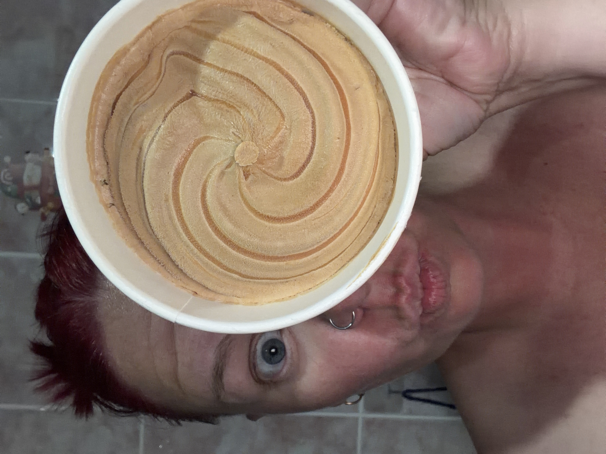 An individual with short, spiky red hair is depicted in a close-up portrait, holding a white cup in their right hand, partially obscuring the right side of their face. The visible left eye is wide open with a dilated pupil, and the mouth is pursed into a "kissing" pose. A silver hoop nose ring is present on the left nostril, and a gold hoop earring is visible on the left earlobe. The cup contains a light brown substance with a distinct, concentric spiral pattern imprinted on its surface. The background consists of light-colored tiled walls, indicative of an indoor environment, possibly a bathroom. An indistinct decorative object is partially visible in the upper right background. The image contains no discernible elements to indicate a specific geographic location such as Chaikovsky, Russia.