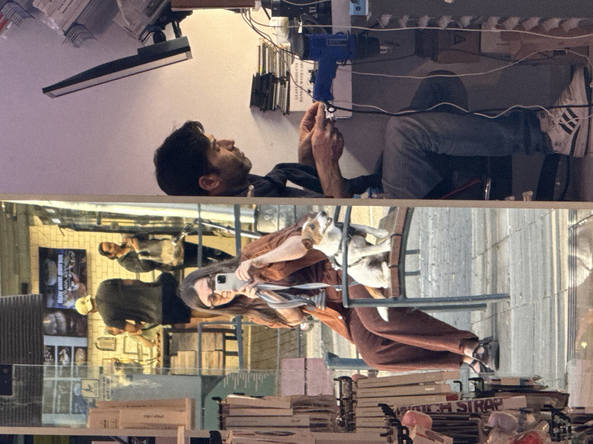 The image depicts a man sitting and working on what appears to be a repair or crafting project in an indoor setting. His surroundings include a desk or workbench with various tools, equipment, and materials. The man is wearing jeans and a shirt, and he is focused on his work. On the other side of the glass, there is a woman taking a picture with a cell phone of herself and a dog sitting in a chair, while another person can be seen in the background behind her. Behind the woman are shelves filled with what appear to be movie posters and books. The location is in Raanana, Israel, as evidenced by the storefront window and products visible in the scene.