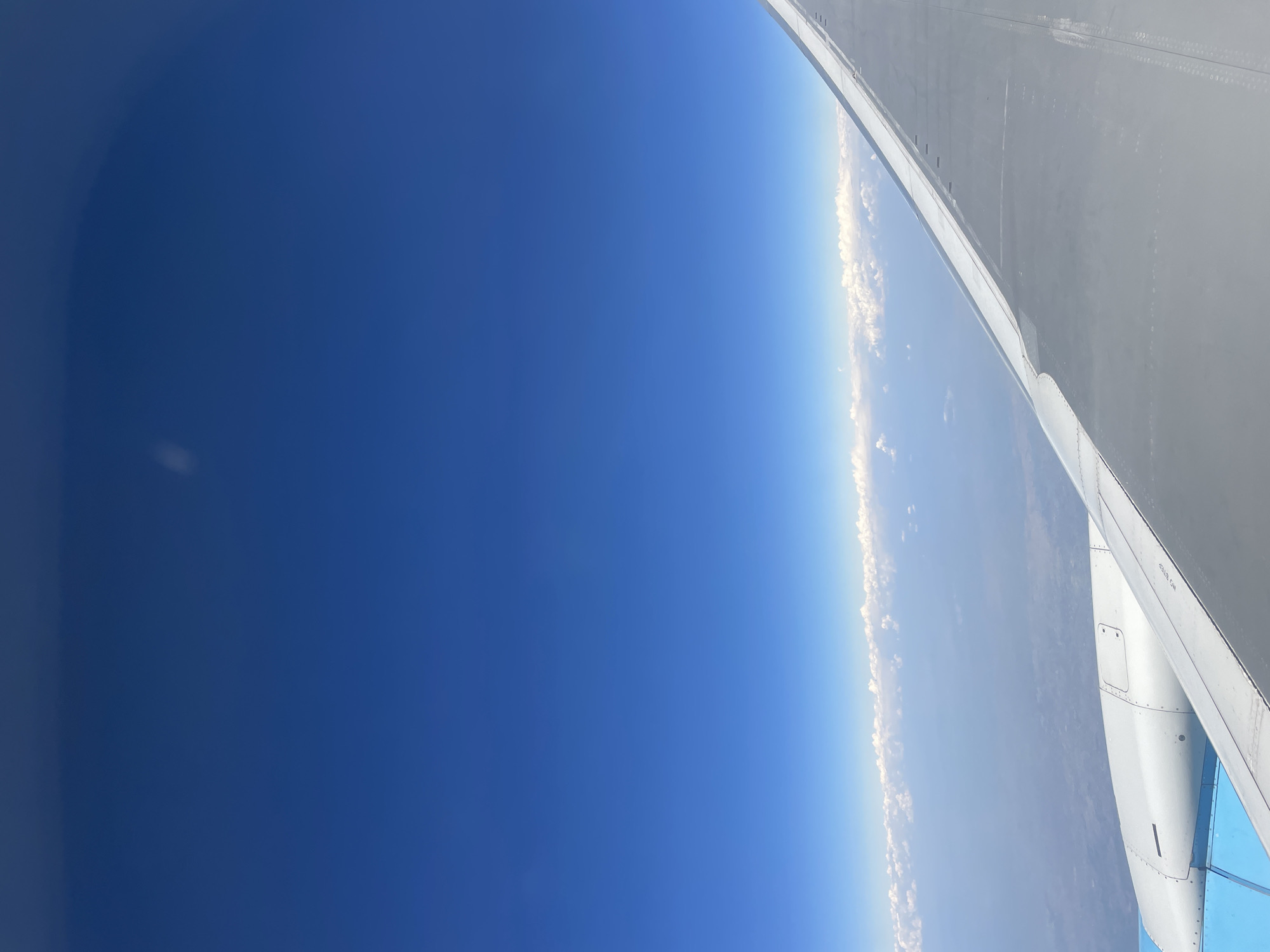 The image shows a view from an airplane window. The wing of the plane is visible on the right side of the frame. The sky is mostly blue, with some clouds near the horizon. The ground below is visible through the clouds. The location is not discernible from this image.
