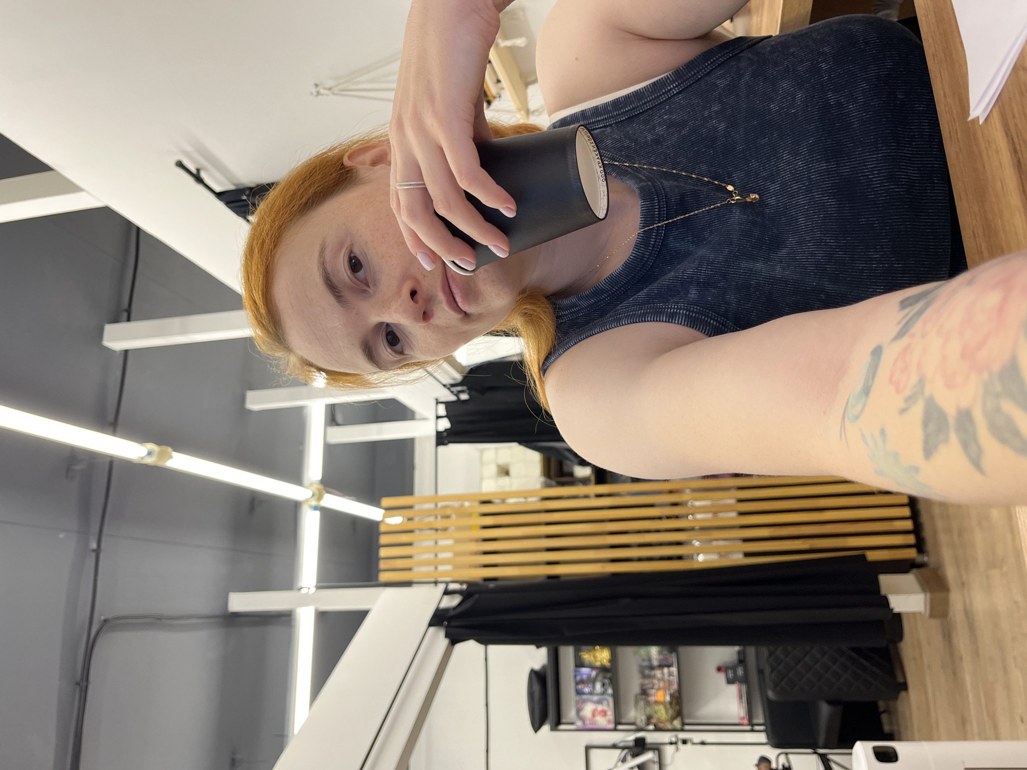 A woman with red hair is holding a black cup and looking at the camera in an indoor setting. She is wearing a dark blue sleeveless top and a gold necklace. There is a tattoo on her left arm. In the background, there are shelves with items, black curtains, a wooden divider, and fluorescent lighting on the ceiling. The floor is made of wood. The scene appears to be a commercial or service space, possibly a shop or salon, in Chaikovsky, Russia.