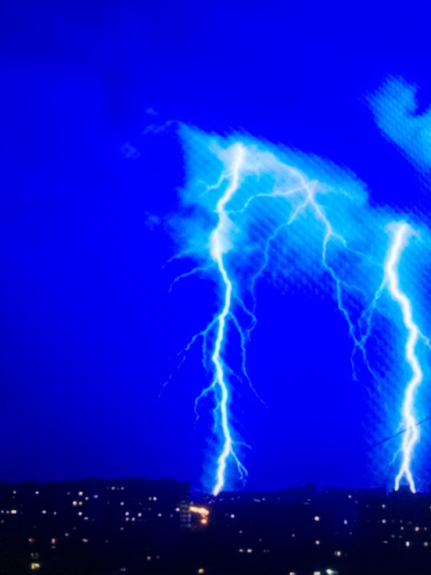 A nighttime scene captures multiple lightning strikes illuminating the deep blue sky over a cityscape. The lightning forks are bright white and branch out across the upper portion of the image. The city below is dark, with scattered points of light from buildings. The image appears to be taken from a distance, with the cityscape forming a dark silhouette against the illuminated sky. The location is not specified.