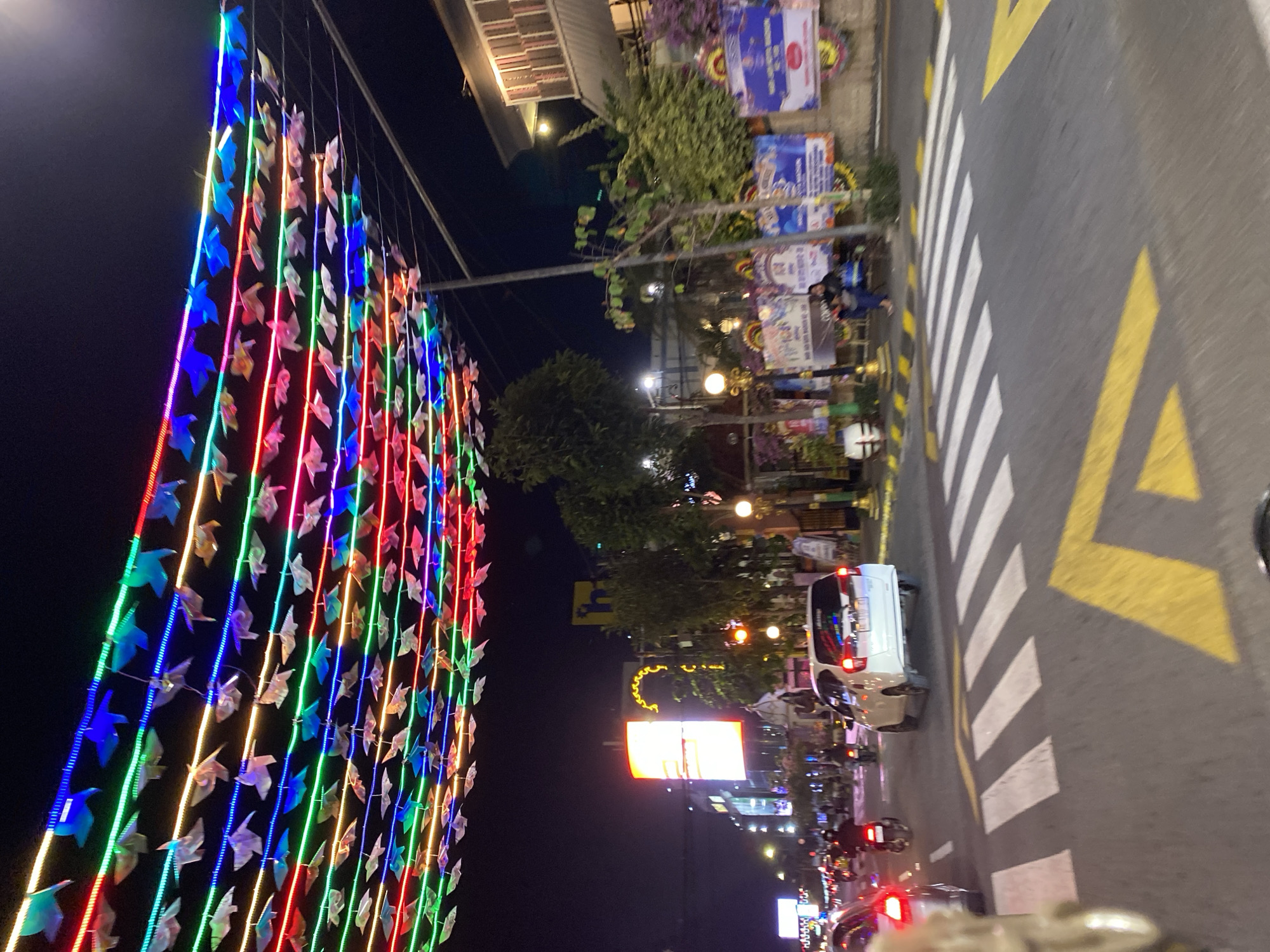 This media file captures a vibrant street scene at night in Madiun, East Java, likely during a festive occasion. The foreground is dominated by a striking display of colorful lights. Rows of small, origami-like figures are strung along wires, illuminated by strings of LED lights in a spectrum of blues, greens, reds, and pinks. These lights create a dazzling, cascading effect, suggesting a celebratory atmosphere.
The scene is set on a street with clear road markings, including a pedestrian crossing with white stripes and yellow directional arrows. The street is paved, and the presence of vehicles indicates it's a public thoroughfare. A white van is visible, parked or moving along the road. Several motorcycles are also present, some with illuminated taillights, adding to the nocturnal energy of the scene.
In the background, buildings line the street, some with awnings and windows. Trees with lush green foliage are interspersed between the structures, softening the urban landscape. Various signs and posters are mounted on the buildings, some featuring text and graphics that are partially visible. One sign displays a yellow, decorative curly motif above a lit rectangular billboard with text. The lighting suggests it is nighttime, with artificial lights illuminating the area. The overall impression is one of a lively, illuminated street, possibly a pedestrianized area or a popular gathering spot during an event. The image evokes a sense of festivity and urban activity after dark.