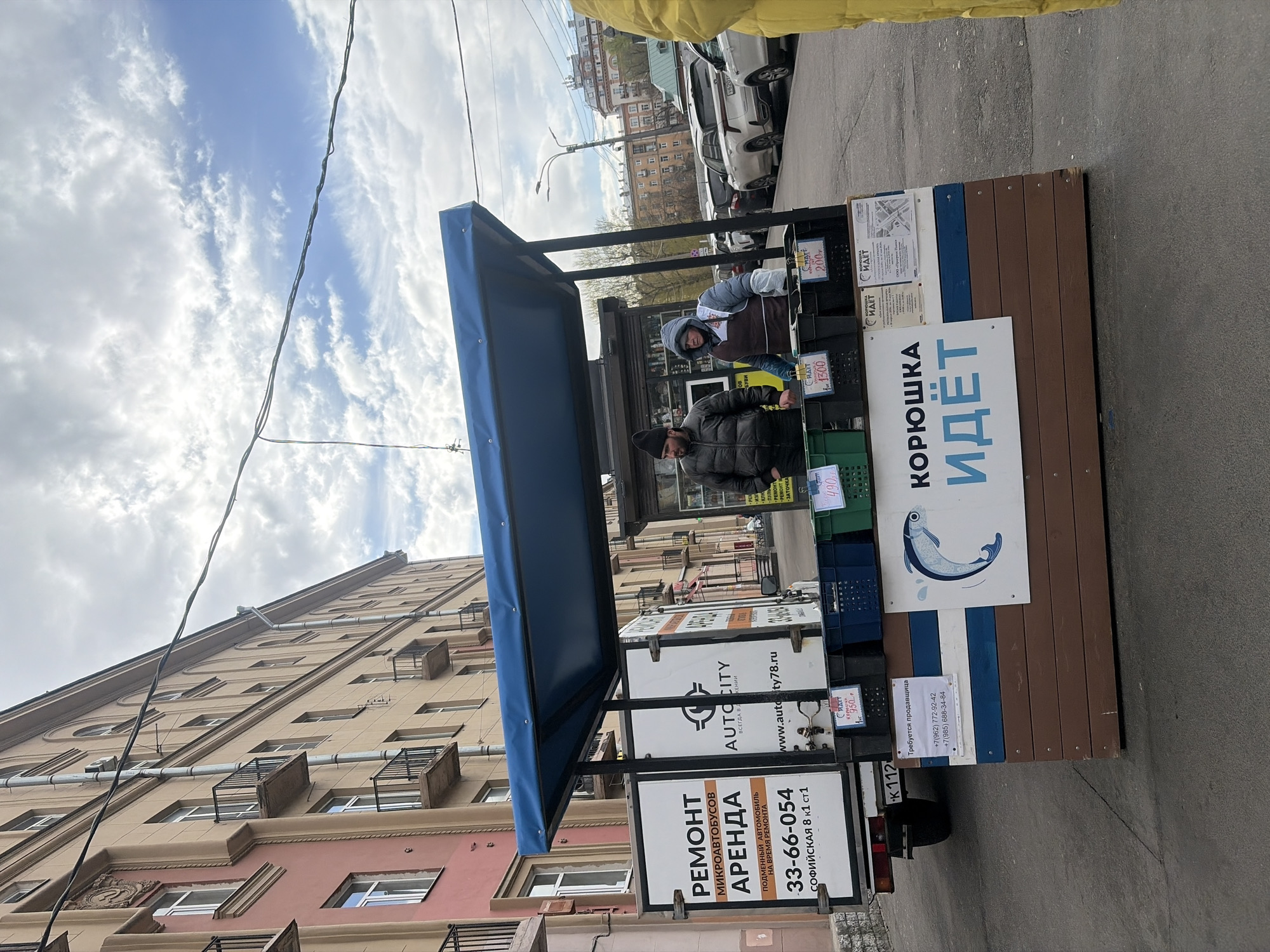 The image shows a street scene with a fish market stall in Saint Petersburg, Russia. Two people are behind the stall. The stall has a blue awning, and signs advertising "Koryushka is here" in Russian (Корюшка идёт), along with contact information and "Auto City" logos. Signs advertising fish prices are displayed. On the side of the vehicle is a sign that reads "MICROBUS REPAIR RENT" "Replaced vehicle during repair" along with a phone number and address: "SOFIYSKAYA 8 k1 ct1". The backdrop includes a multi-story building and other parked cars. The sky above the building is a pale blue, and the streets are paved.