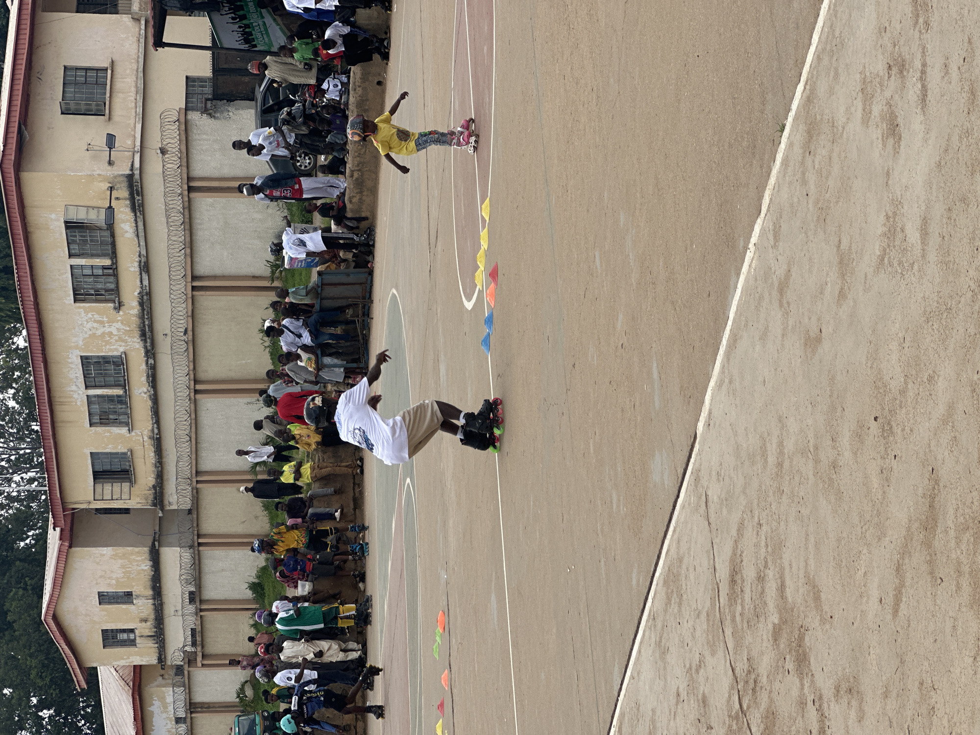In the city of Jos, Nigeria, an outdoor basketball court serves as the venue for an inline skating event. A young boy, clad in a yellow shirt and patterned pants, skates in the foreground, his arms outstretched for balance. He wears a helmet. In the mid-ground, another boy on inline skates, dressed in a white t-shirt and shorts, practices his moves, also sporting a helmet. A line of colorful cones is set up on the court, suggesting a slalom or obstacle course. The background reveals a crowd of spectators, many of whom appear to be children, observing the skating activity. They are gathered near a building complex with a pale yellow facade, barred windows, and a corrugated metal roof. A wire fence with barbed wire runs along the perimeter of the property. The court surface is concrete, marked with white lines.