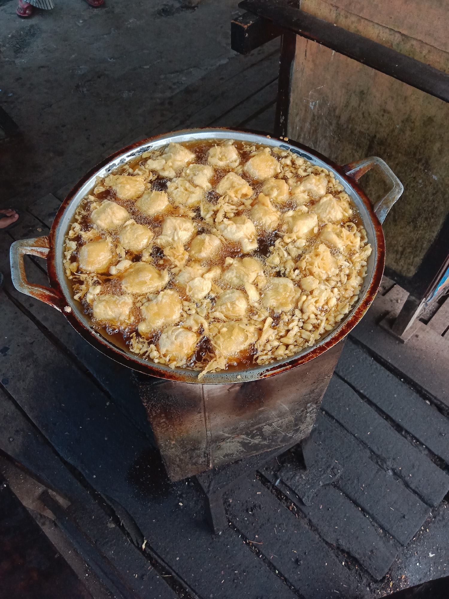 A high-angle shot captures a large, metal basin filled with bubbling oil, in which numerous golden-brown fritters are being fried. The fritters appear to be bite-sized and irregular in shape, some partially submerged and others floating on the surface. The basin itself is worn and slightly rusted, with two prominent metal handles. It rests on a dark, textured surface that resembles a makeshift stove or a platform, possibly constructed from wood and metal. The surrounding environment is rustic, with visible wooden planks and dark, grimy surfaces, suggesting an outdoor or informal setting. In the upper left corner, the feet of at least two individuals are visible, wearing flip-flops, indicating human presence and activity. The overall scene depicts a street food vendor preparing fritters in Pohsangit Leres, Indonesia.