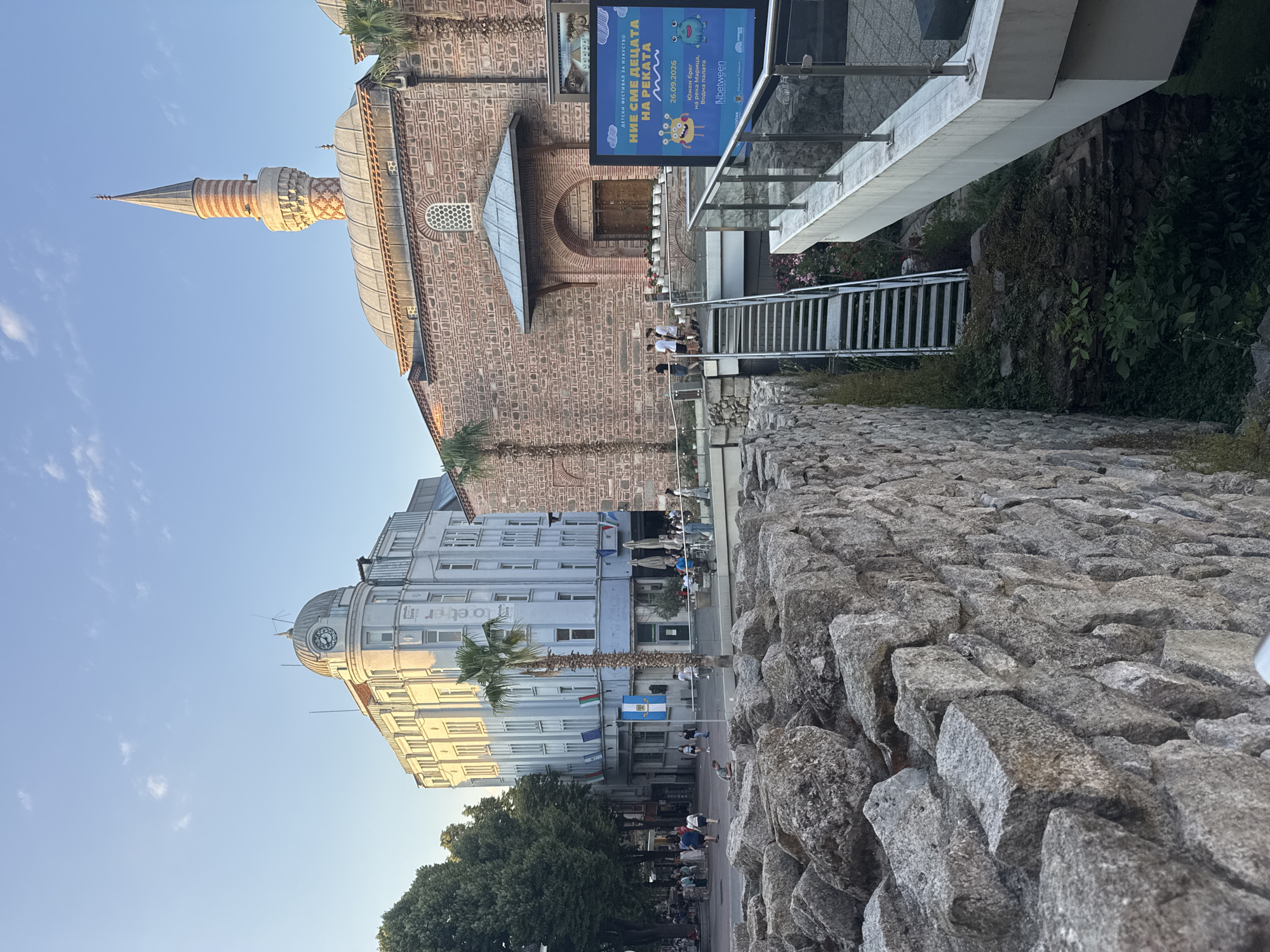 This detailed image captures a vibrant street scene in Plovdiv, Bulgaria, at what appears to be late afternoon or early evening, characterized by warm, golden sunlight illuminating the architecture under a clear blue sky with sparse white clouds.
In the immediate foreground on the left, a substantial, rough-hewn stone wall, likely part of ancient Roman ruins, angles upwards and to the right, forming a textured barrier. Below this wall, a trench reveals further ancient foundations covered in lush green and dark vegetation. A modern, light-grey metal staircase descends into this excavated area from the mid-ground.
The mid-ground features a modern concrete walkway with glass and metal railings, extending from the right towards the center. On this walkway, a blue billboard displays colorful cartoon monsters and text in Bulgarian, announcing a "Children's Art Festival" on "26.09.2026" and mentioning "Община Пловдив" (Municipality of Plovdiv). Behind the billboard and walkway stands the Djumaia Mosque (Cuma Camii), a historical structure built of reddish-brown brick with a gabled roof and a central dome. Its prominent minaret rises tall with alternating horizontal bands of reddish-brown and lighter stone, culminating in a pointed spire. An arched wooden entrance with several white potted plants sits beneath a small protective roof on the mosque's side. Several palm trees are scattered around the mosque and walkway area. Small groups of people are visible on the walkway and descending the stairs, casually dressed and observing the surroundings.
In the background on the left, a multi-story, light-colored building with classical architectural elements dominates the skyline. This structure features a prominent domed clock tower at its corner, adorned with a decorative railing and a small antenna. Below the clock tower, a section of the building displays hanging flags, including the Bulgarian and European Union flags. In front of this building, a bustling pedestrian street is visible, lined with mature green trees. Numerous individuals, dressed in casual attire, walk along the street, some gathered near outdoor seating areas suggesting cafes or restaurants.
The composition juxtaposes ancient ruins, historical architecture, and modern urban elements, all under the soft glow of the setting sun, firmly situating the scene in the historical and vibrant center of Plovdiv, Bulgaria.