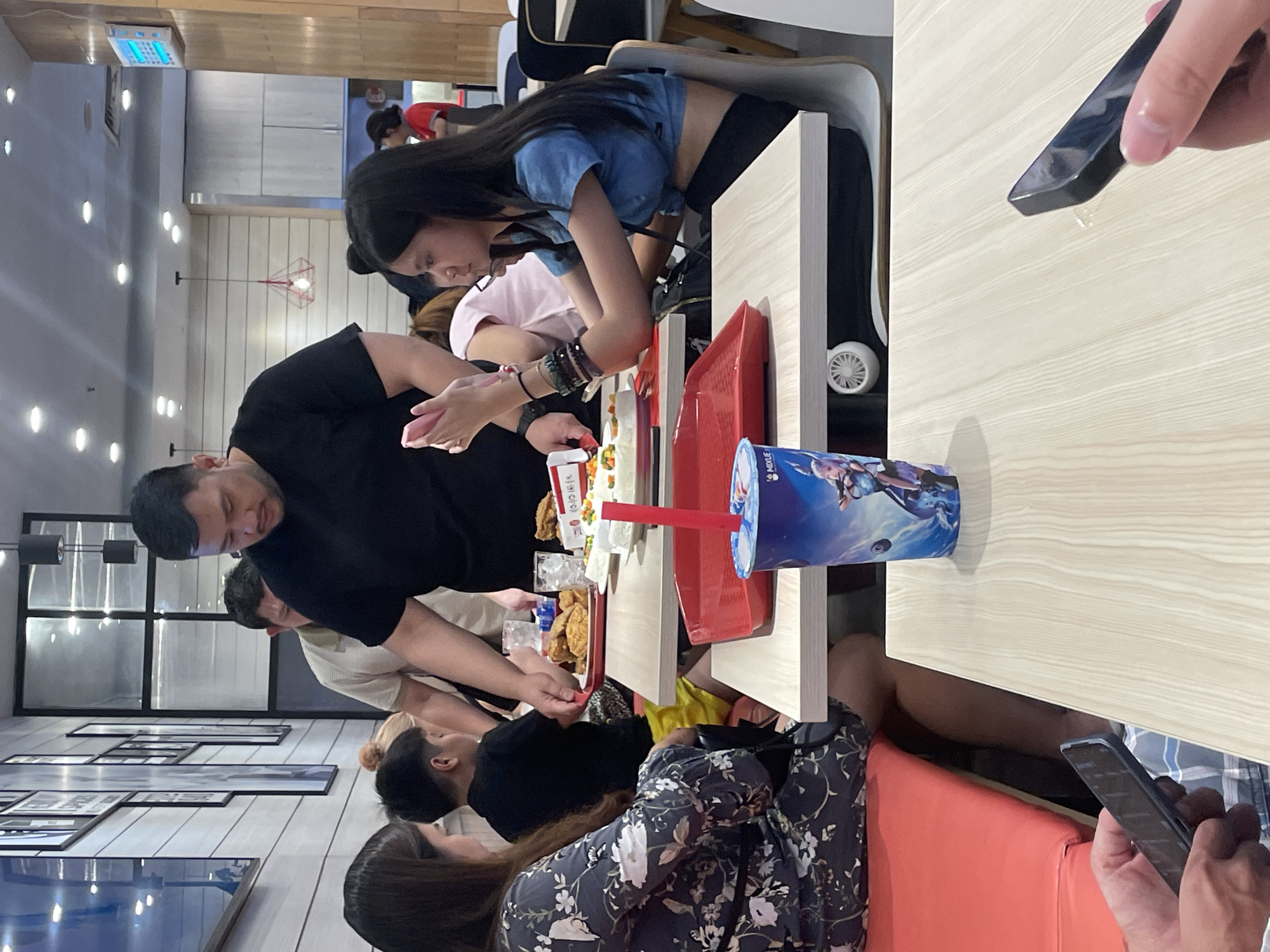 The image depicts a group of people at a fast-food restaurant in Đà Nẵng, Vietnam. In the foreground, a person's hand holds a smartphone over a light-wood table. To the left, another person, partially visible, holds a smartphone. A blue cup with an anime-style character on it sits on the table. Behind the cup, a red tray holds food, possibly fried chicken and rice. A man in a black t-shirt is standing, holding another tray of food, and appears to be serving the people seated. Several other individuals are visible, including a woman in a floral dress and a young boy wearing yellow shorts. The restaurant interior features light-colored walls with framed pictures, modern lighting fixtures, and other tables and chairs. The scene suggests a casual dining experience.