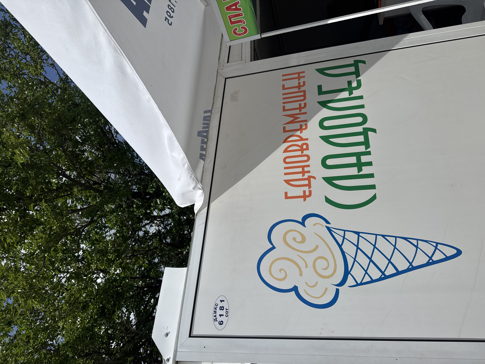 A street-level shot shows an advertisement for "СЛАДОЛЕД", or "Ice Cream", in Pesnopoy, Bulgaria. The advertisement is located on the side of a white vendor cart or building. The image depicts an ice cream cone in light yellow with blue outline, along with Cyrillic script in green and orange. The script translates to "Sweet Ice Cream for Everyone". A white awning partially covers the top of the image, and the top of a green sign saying "Сла..." can be seen as well. Some trees are visible to the left of the cart. The lower-left side of the vendor cart has a blue label with the number "6181".