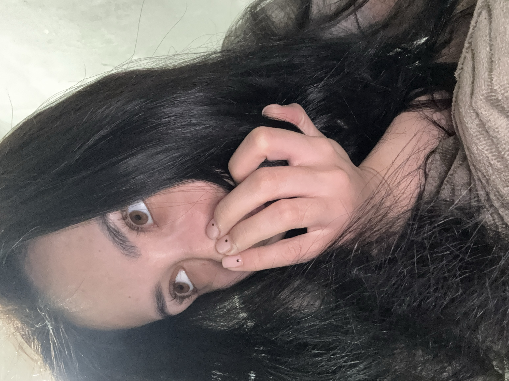 A selfie-style photograph shows a person with dark, straight hair and light brown eyes. The person is fair-skinned. Their face is tilted towards the camera, with a hand near their face; the fingernails are light in color and have black dots. The person is lying on their side. A light-colored, textured fabric can be seen in the upper right corner of the frame. The background appears to be a neutral, light surface. The location of the photograph in Votkinsk, Russia, cannot be verified with the information provided.