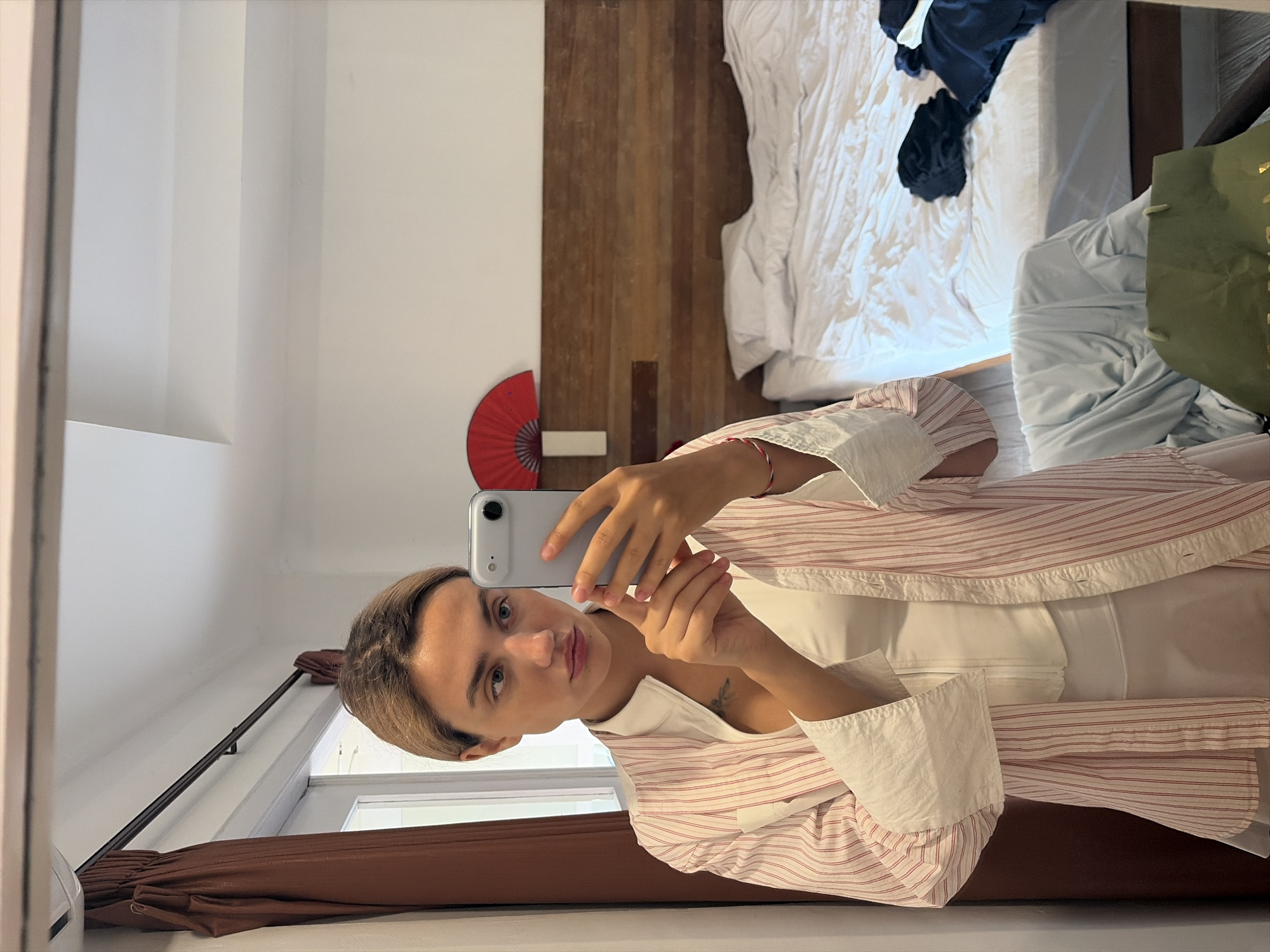 A young woman with blonde hair pulled back is taking a selfie in a mirror. She is wearing a white corset and a loosely buttoned white and pink striped shirt. The room appears to be a bedroom with a made bed, a wooden headboard, and a red fan decoration on the wall. A brown curtain hangs on the left side of the frame, and a window is visible behind it. A green shopping bag is visible in the foreground. The scene is likely in a hotel room in North Kuta, Indonesia.