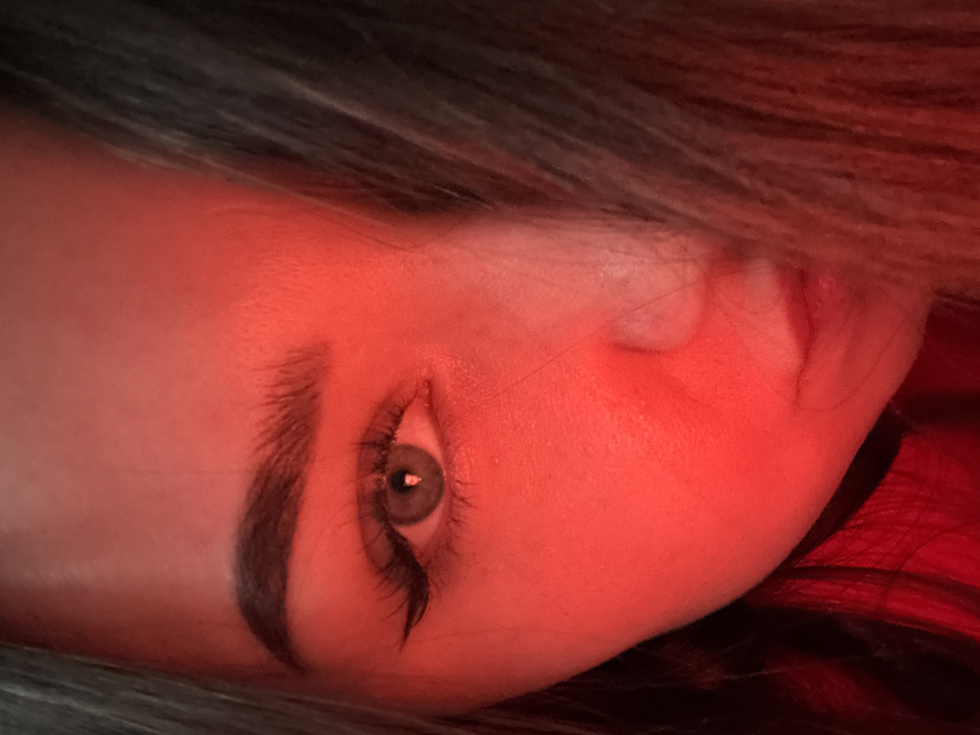 The image presents a close-up portrait of a person, likely female, under intense red or reddish-orange lighting. The subject's head is rotated approximately 90 degrees counter-clockwise, with dark hair visible along the top and bottom edges of the frame.
The central focus is on one eye, which has a distinct light-colored iris, possibly green or hazel, contrasting sharply with the dominant red illumination. Long, dark eyelashes frame the eye, and a well-defined, dark eyebrow arches above it. The skin of the forehead and cheek glows with a vibrant red hue due to the lighting, obscuring natural skin tones but highlighting contours. A portion of the nose and mouth can be faintly discerned on the right side of the face, also bathed in the red light.
The dark hair appears to be long and flows around the head, offering a darker backdrop to the brightly lit face. No specific activity is occurring beyond the subject posing for the photograph. The expression conveyed by the visible eye is steady and neutral. The strong red lighting creates a dramatic and atmospheric mood, suggesting an indoor setting or controlled light environment. No text is visible in the media file.