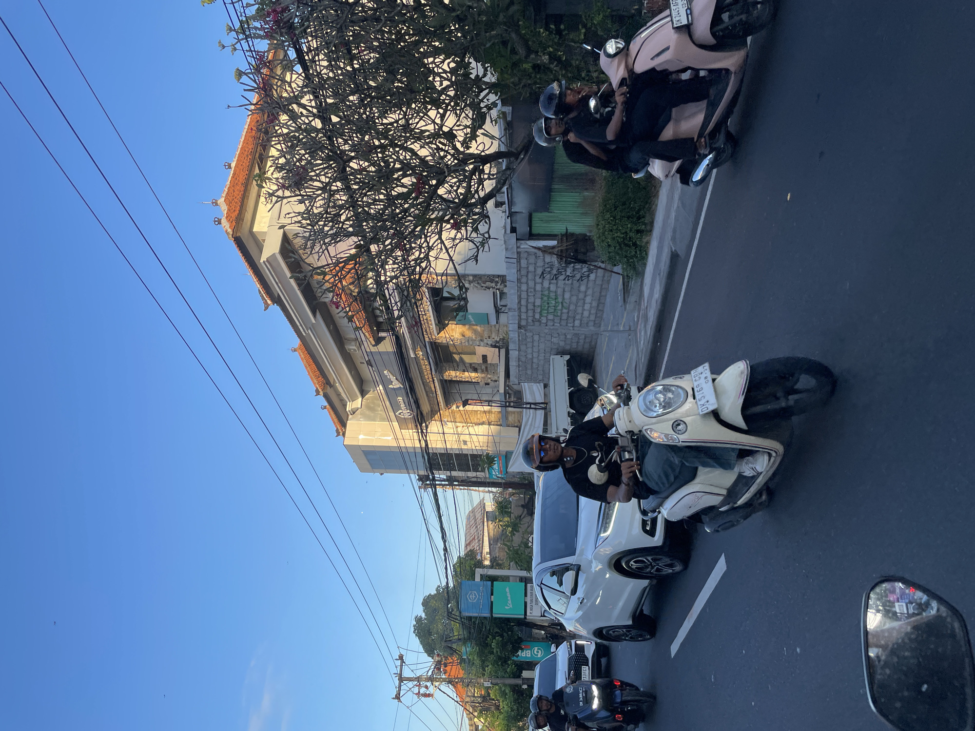 In Denpasar, Indonesia, on a paved road, a motorcade of motorcycles and a white car moves under a bright blue sky. The lead motorcycle, a cream-colored scooter, is ridden by a person wearing a black t-shirt, sunglasses, and a helmet, their legs clad in green pants and white sneakers. Behind them, a pink scooter carries two individuals also wearing helmets. To the left, a white SUV is followed by another motorcycle. In the background, a building with an orange-tiled roof and stone facade stands tall, adorned with a tree displaying bare branches. Utility poles with numerous wires are visible on the left side of the street. The road is marked with white lines, and a traffic mirror reflects a blurry image in the foreground.