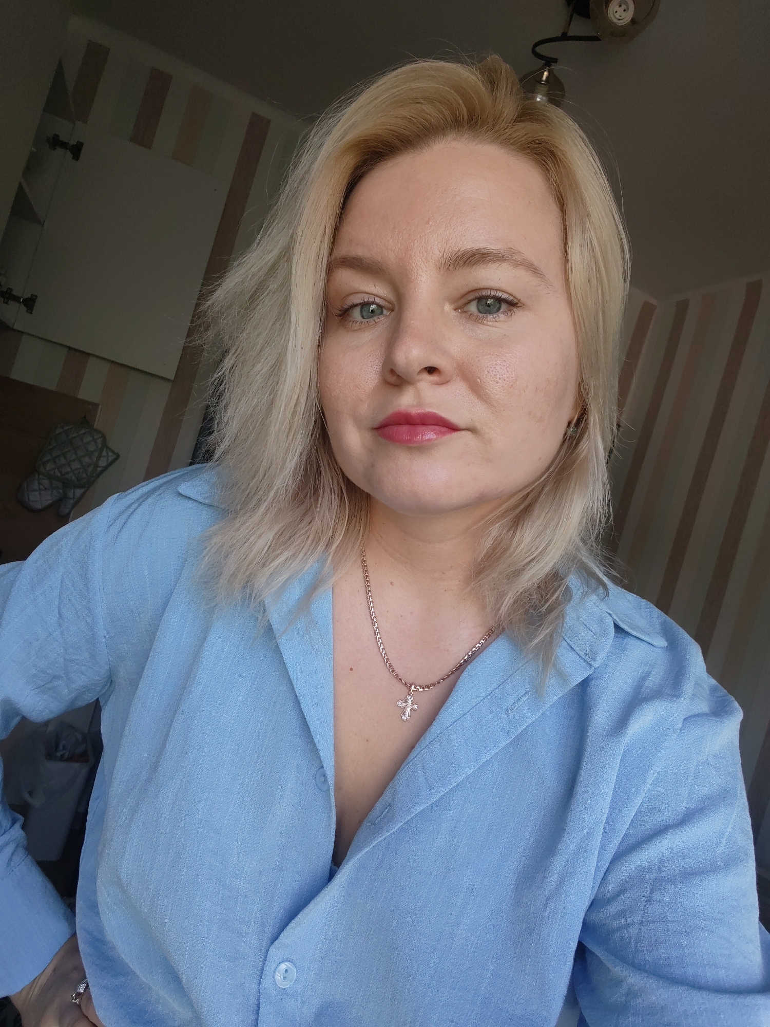 A woman with blonde hair and blue eyes, wearing a light blue button-down shirt and a gold cross necklace, is shown from the chest up, looking directly at the camera. Her makeup includes pink lipstick. The background features a wall with vertical striped wallpaper and a portion of a white cabinet. The lighting suggests an indoor setting, possibly a home in Izhevsk, Russia. The woman has her left hand on her hip, and a diamond ring is visible on her finger.