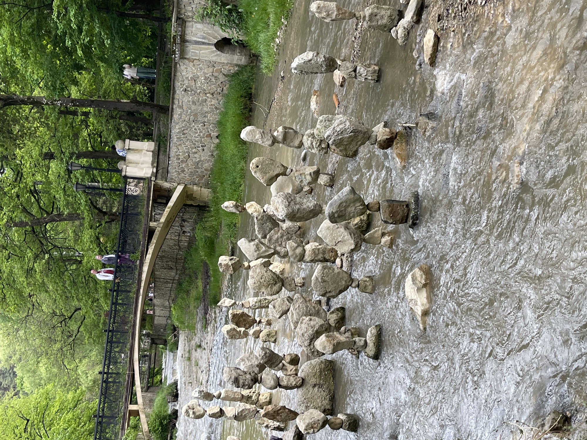 The scene is a stream in Kislovodsk, Russia. Multiple stacks of rocks are balanced in the stream. A stone wall with an archway runs along the right side of the stream. On the left is a bridge with pedestrians crossing it. Trees line the left side. More people are visible in the background. The stream flows from left to right.
