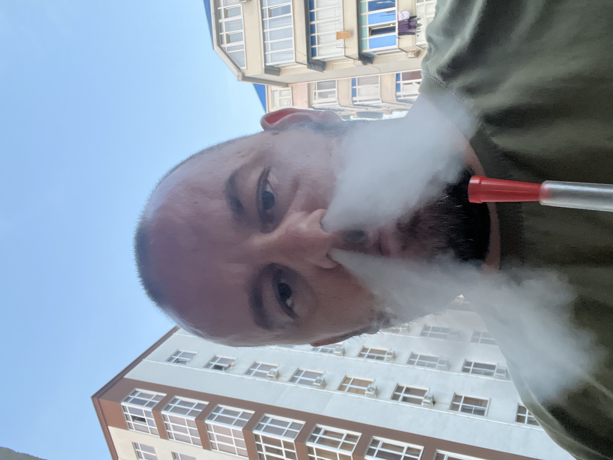 A close-up, low-angle view captures a male subject exhaling a dense cloud of white vapor or smoke from his mouth and nostrils. The man has a shaved cranium and a beard, looking directly into the camera. An olive green shirt is visible on his torso. A red-tipped, transparent hookah hose mouthpiece is positioned near his chin, centered at the bottom of the frame.
The background depicts an urban residential environment under a clear blue sky. To the subject's left, a tall, light-colored building with prominent brown corner detailing and window frames is visible, featuring numerous rectangular windows and several external air conditioning units. To the right, another complex of buildings appears, including a beige building with a blue roof and multiple windows, some exhibiting a blue tint. Laundry is observed hanging from a window on a building in the far right background. The scene is consistent with an urban area during daytime, specifically within the city of Sochi, Russia.