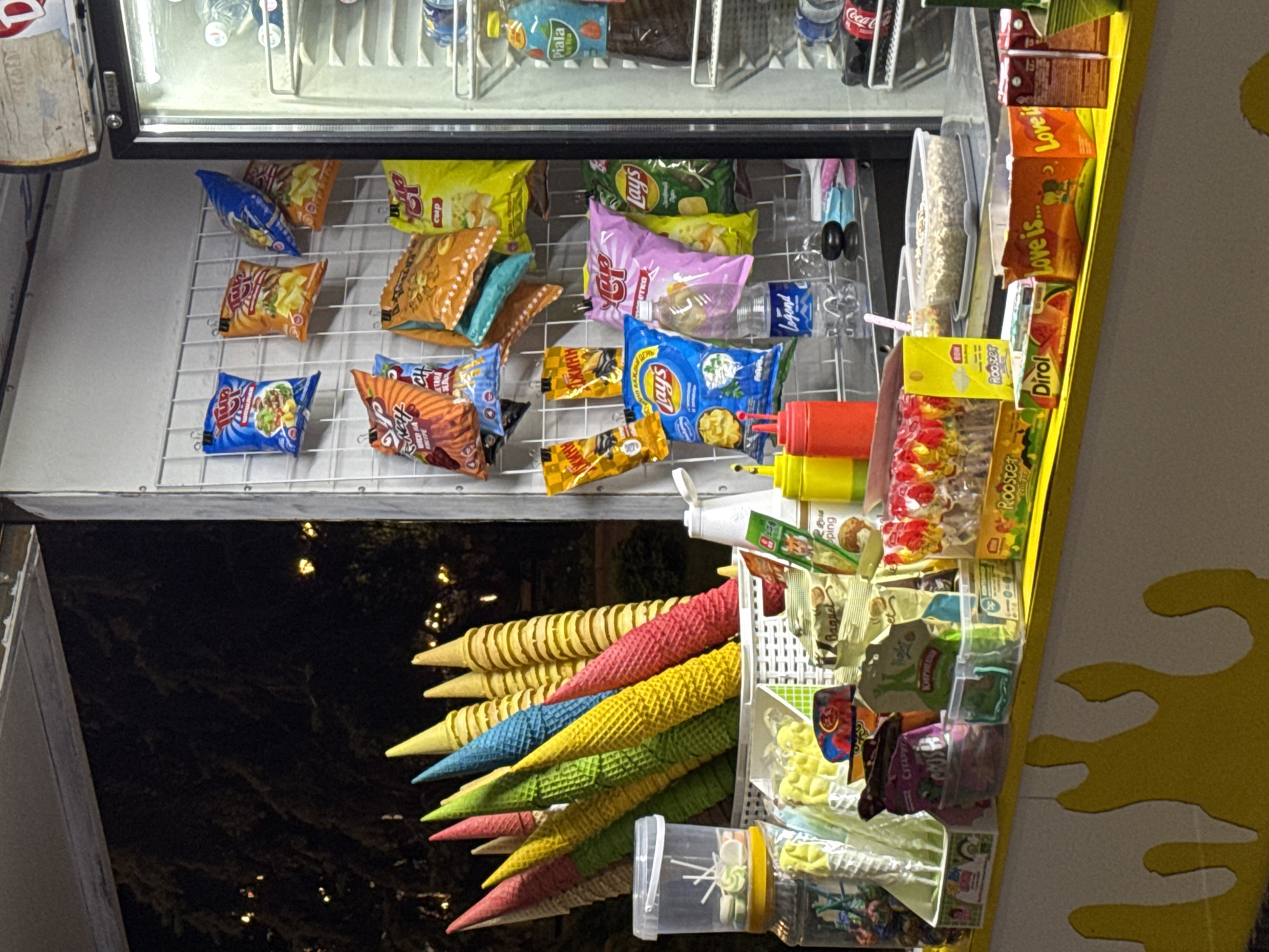 This image shows a street food stall in Bishkek City, Kyrgyzstan, at night. The stall is filled with various snacks and drinks. In the foreground, there are colorful ice cream cones, bags of chips, and packaged candies. A refrigerator to the right is stocked with bottles of water and soda. The background shows trees and lights, indicating an outdoor urban setting. The stall appears to be open for business, with goods displayed for sale. No people are visible in the image.