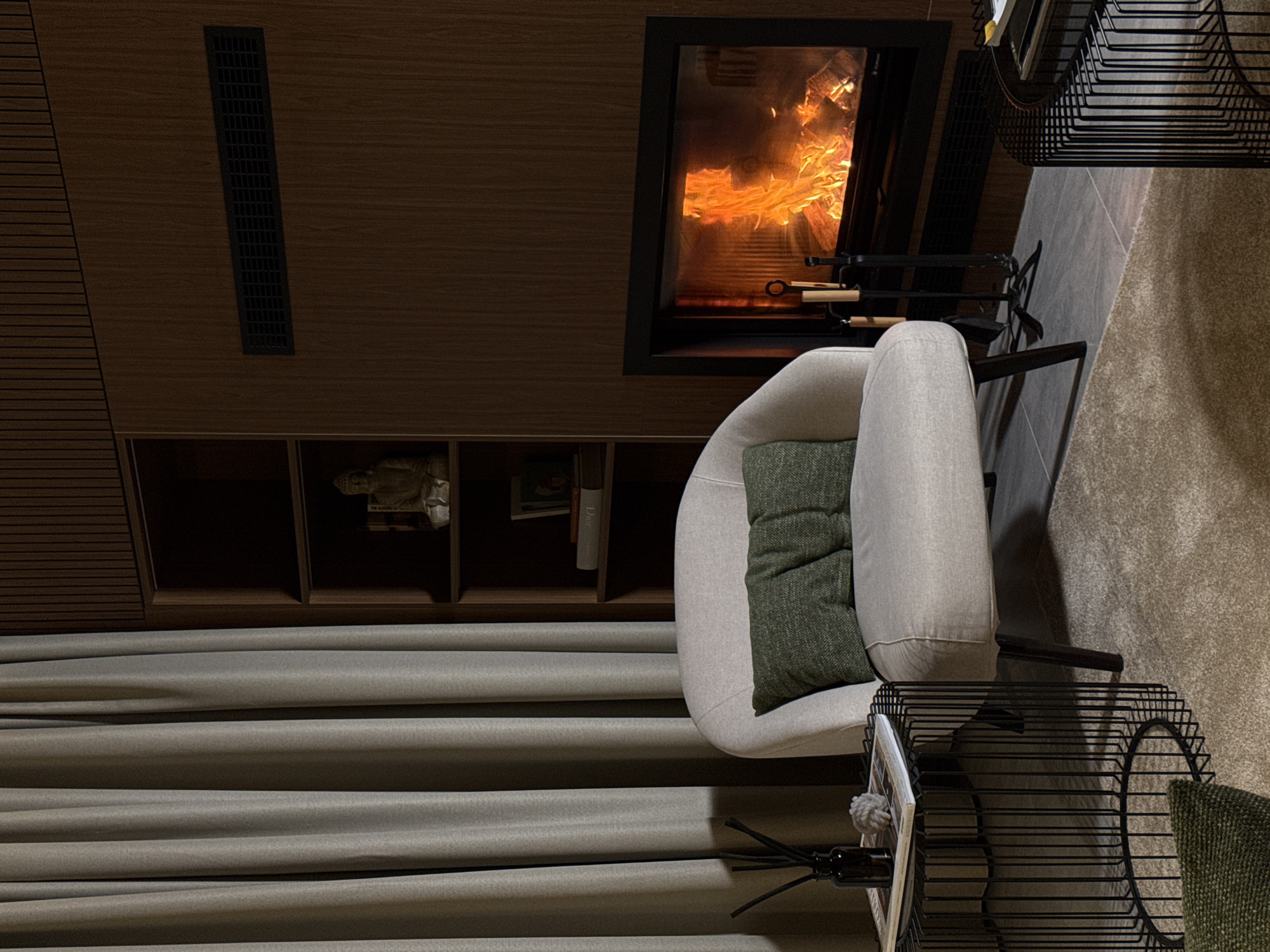 The image depicts a cozy living room scene in Bakovići, Montenegro. A plush, light-colored armchair with two green throw pillows sits on a textured beige carpet. To the left of the armchair is a black wireframe side table with a dark glass diffuser bottle and a book. In the background, a fireplace with a roaring fire is set into a dark wood-paneled wall. Above the fireplace is a horizontal vent. To the left of the fireplace is a dark wood shelving unit containing a small Buddha statue and books. A tall, light gray curtain hangs on the far left side of the image. The overall ambiance is warm and inviting.