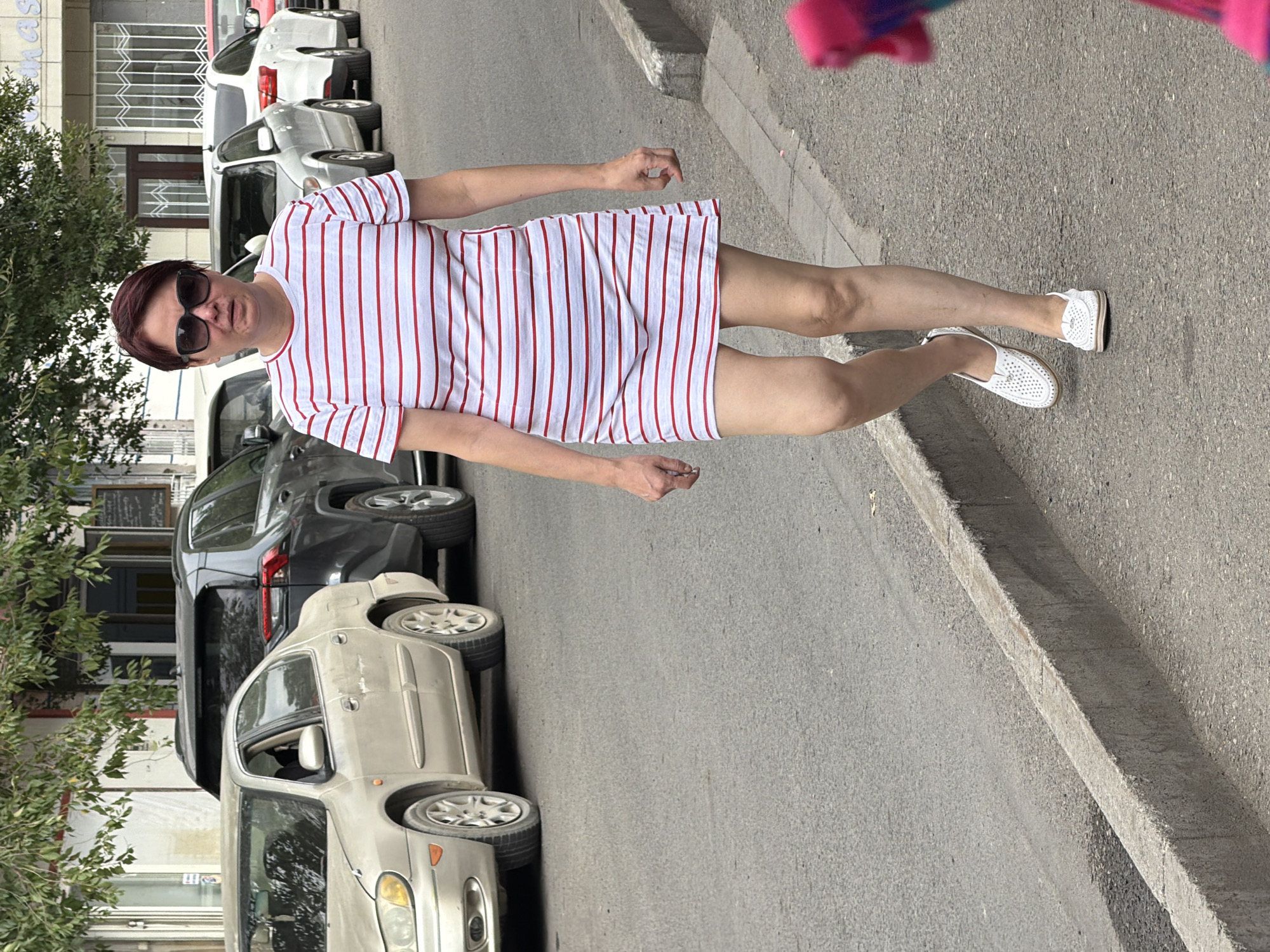 In an outdoor urban setting, a woman with short, dark red hair and sunglasses walks on a paved street. She is wearing a white and red horizontally striped, short-sleeved dress and white perforated flat shoes. The woman is walking towards the viewer on the right side of the frame. Behind her, several cars are parked along the street, including a light-colored sedan and a dark-colored SUV. Buildings with windows and awnings line the street in the background, and trees with green foliage are visible above the buildings. The street is bordered by a concrete curb.