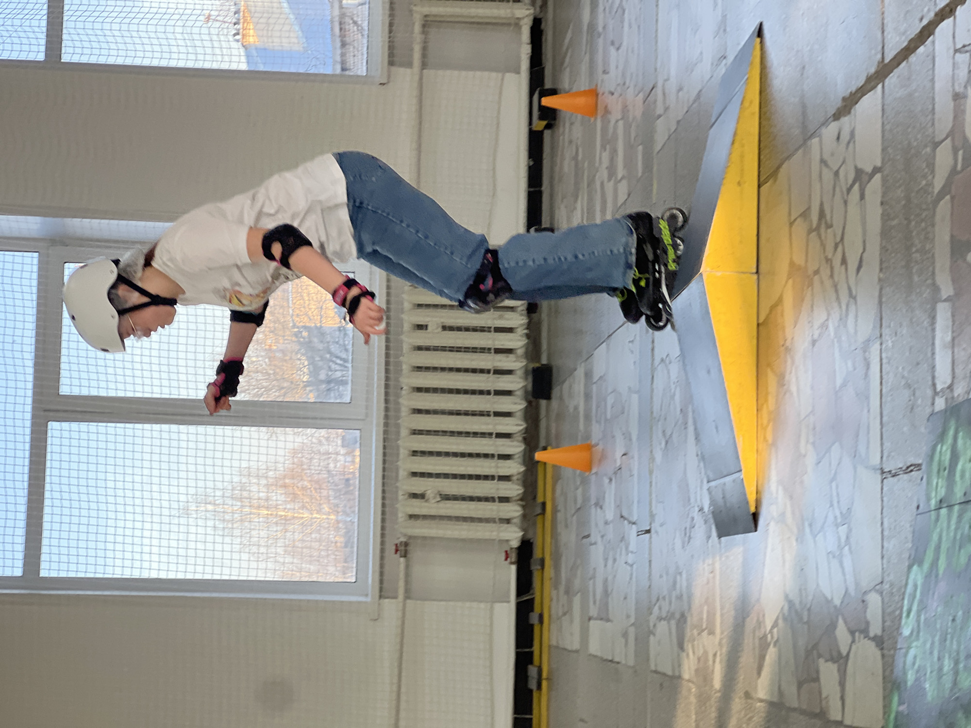 A person wearing a white helmet, white t-shirt, blue jeans, and black inline skates is riding a skateboard ramp indoors. The person is wearing black elbow and wrist pads. The ramp is painted gray and yellow. Orange cones are placed near the ramp. The location appears to be an indoor skate park or recreational facility in Chaikovsky, Russia. Windows and a radiator are visible in the background. The floor is tiled and the walls are painted white.