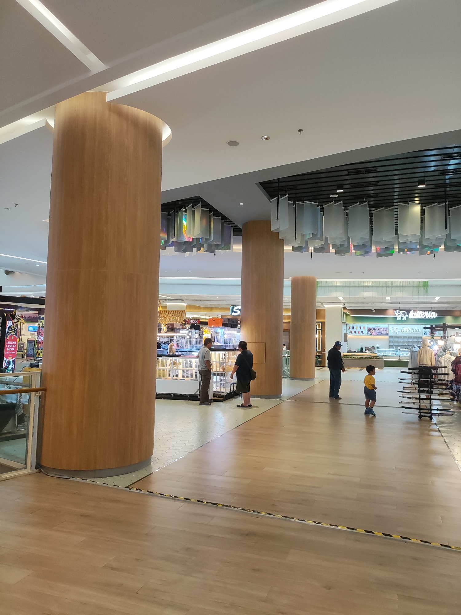 The image depicts an indoor scene, likely a shopping mall, with a polished wooden floor and large, cylindrical wooden columns. In the background, there are retail displays and people, suggesting a commercial environment. One prominent sign reads "batteria" with a stylized elephant logo. There are several individuals present: a man in a gray shirt and brown pants, a woman in black, a man in a dark jacket and blue jeans, and a young boy in a yellow shirt and blue shorts. The ceiling features modern lighting fixtures and suspended decorative elements. The scene appears to be in Lambangjaya, Indonesia, as indicated by the location information provided. The presence of social distancing markers on the floor, visible as yellow and black tape, suggests the photo was taken during a period when such measures were in effect.