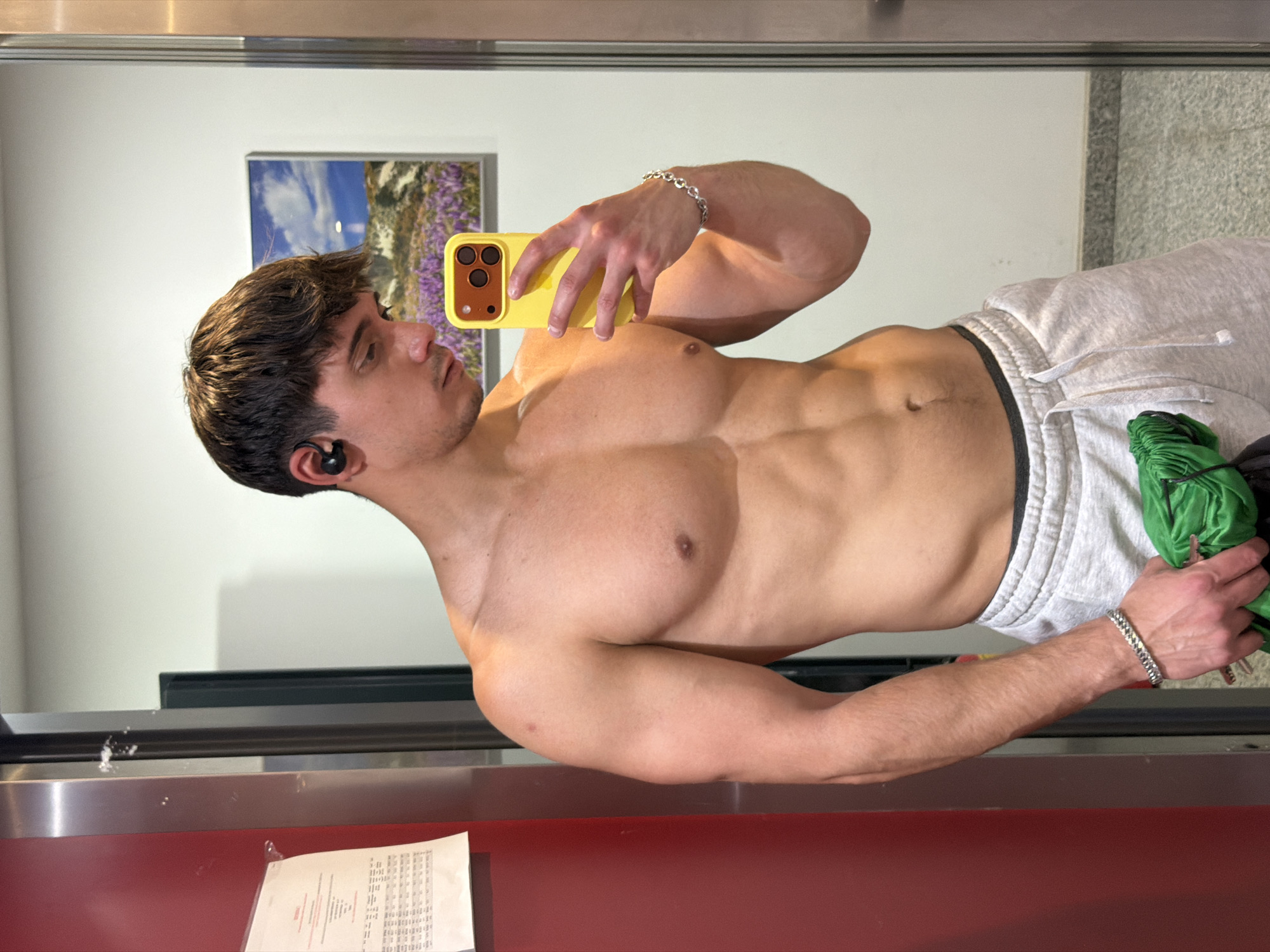 A shirtless man with brown hair and an earbud in his left ear is taking a selfie with a yellow cell phone in front of a mirror. He is wearing light gray sweatpants and a silver bracelet on his left wrist. In his right hand, he holds a green object. Behind him in the mirror, a picture hangs on the wall. The location is not specified, but could be in Sofia, Bulgaria. A document is on a shelf in the foreground.