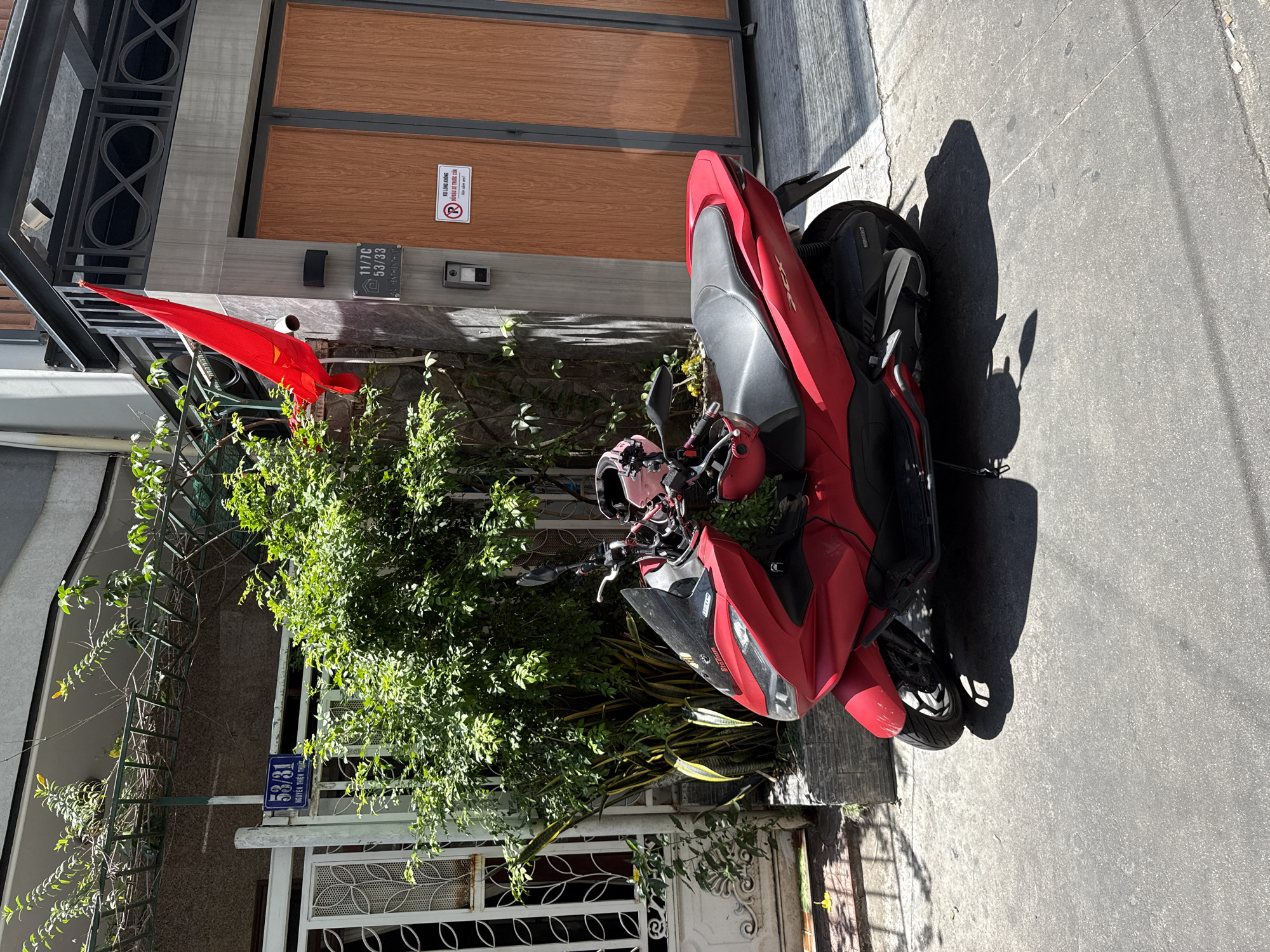 The image shows a red scooter parked on the sidewalk in front of a building. A Vietnamese flag hangs from the building's exterior. The building's address is 53/31 Nguyễn Thiện Thuật, Nha Trang Ward, Vietnam. Next to the scooter is a patch of greenery obscuring the building's door. Another address, 11/7C 53/33 Nguyễn Thiện Thuật, Nha Trang Ward, Vietnam, is visible next to the wooden door. There is a sign reading "VUI LÒNG KHÔNG ĐỔ ĐẦU XE TRƯỚC CỦA" on the wooden door.
