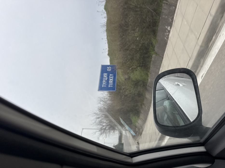 The image depicts a roadside scene captured from within a vehicle, likely located near the Lesovo border crossing in Bulgaria, indicating proximity to Turkey. A blue rectangular road sign, prominently positioned on an embankment to the right, displays "ТУРЦИЯ" (TURKEY) above "TURKEY," followed by "0.5," signifying 0.5 kilometers to the Turkish border. The sky is overcast and uniformly grey. The foreground shows a portion of the vehicle's white interior door frame and a side mirror. The mirror's reflection reveals the occupant of the vehicle, wearing a light green shirt. To the left, a paved road is visible, curving slightly, with several bare-branched trees and streetlights in the distance. The area adjacent to the road on the right features a concrete sidewalk or shoulder, bordering a gentle slope covered with dry bushes and some green vegetation.