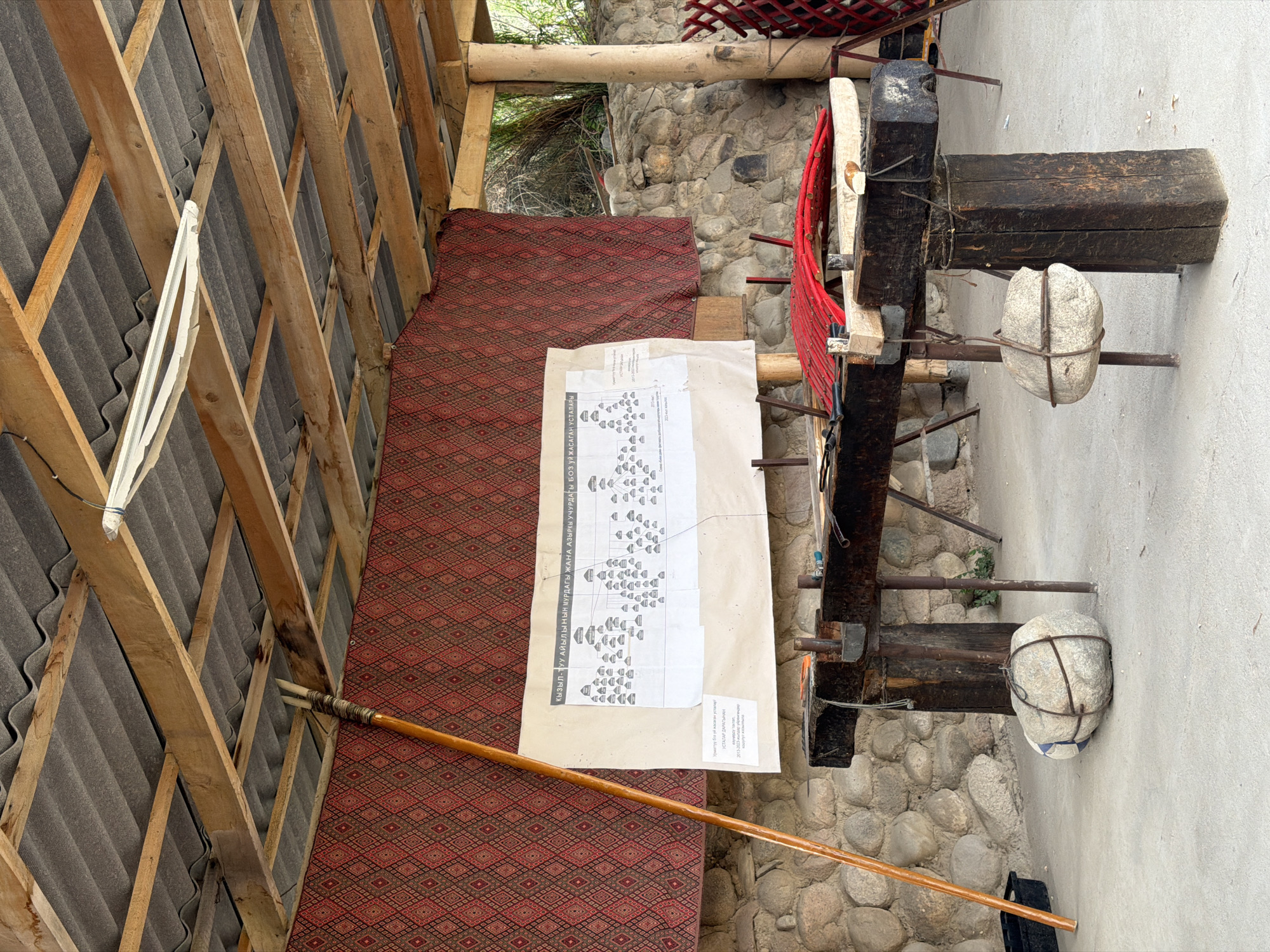 This image captures an outdoor workshop or exhibition space in the city of Кызыл-Туу, Kyrgyzstan. The scene is set under a rustic wooden roof supported by beams, with corrugated roofing material overhead. A patterned red textile hangs as a backdrop.
Dominating the center of the image is a large, hand-drawn diagram or chart displayed on a piece of paper, possibly a family tree or organizational chart, with rows of small, house-like icons connected by lines. The text above and below the chart appears to be in a Cyrillic-based script.
In front of the chart is a sturdy, heavy wooden workbench. This workbench is supported by substantial wooden legs and appears to be a functional piece of equipment for craft or trade. Suspended from the workbench are two large, round stones, each wrapped with wire or rope for stabilization and potentially for use as weights or counterbalances. A long, wooden pole, possibly a tool or staff, leans against the left side of the workbench.
The environment suggests a rural or traditional setting, with a stone wall visible behind the workbench and some greenery in the background. The ground is concrete. Red materials are also visible on the workbench, suggesting ongoing work or display. There are no people visible in the frame.