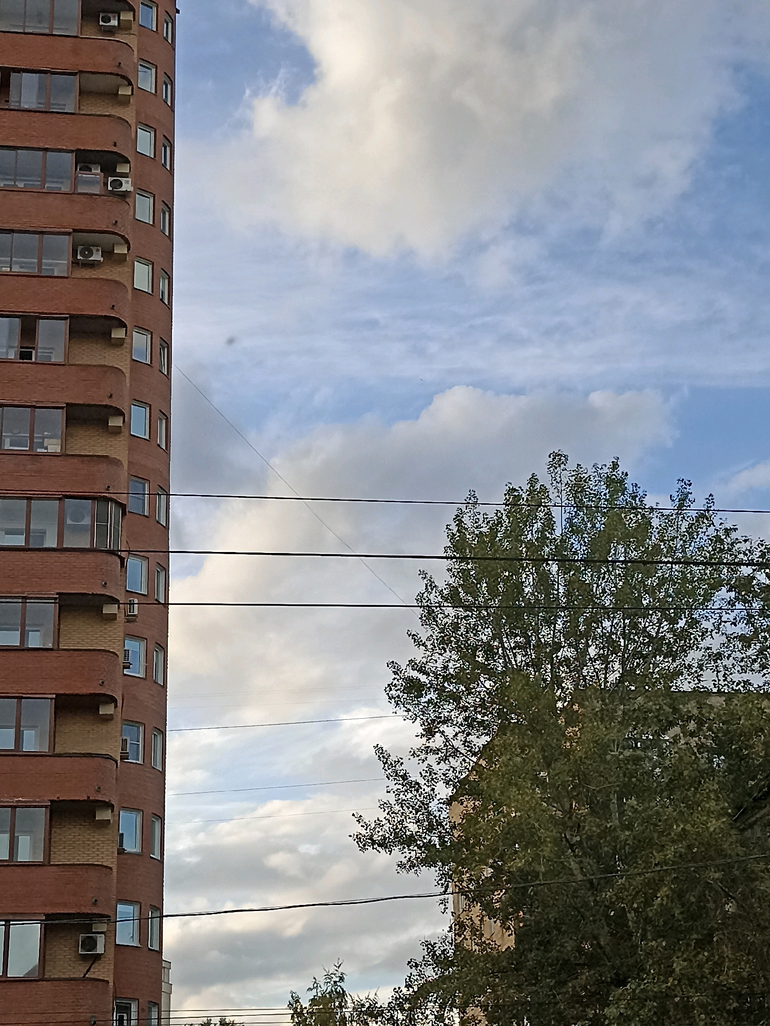 A tall, curved building made of red brick and yellow accents stands on the left side of the frame. It has multiple stories with many windows, some of which appear to be French windows with balconies. Air conditioning units are visible on some of the balconies. In the background, the sky is partly cloudy with white and gray clouds against a blue sky. A large tree with green leaves dominates the right side of the image, partially obscuring another building behind it. Several power lines run horizontally across the frame, in front of both the building and the tree. The scene appears to be an outdoor urban environment.