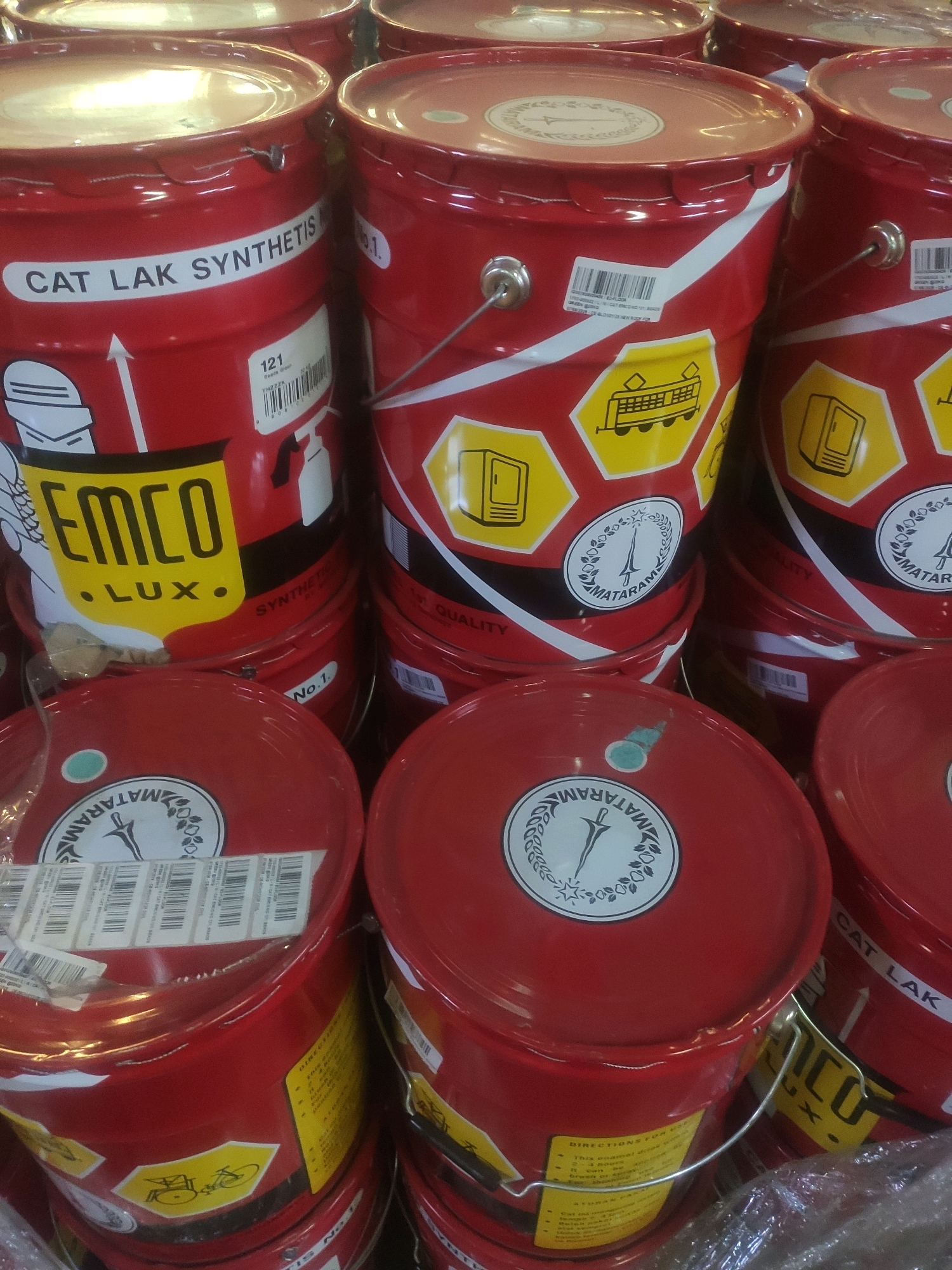 The image displays a stacked arrangement of red metal buckets, likely containing paint or industrial fluids. The buckets are adorned with white and yellow labels and graphics. Some labels feature text such as "CAT LAK SYNTHETIS", "EMCO", and "LUX.". Other labels show icons like a bicycle and a train car. Several buckets also have white circular emblems with the word "MATARAM" in a semicircle. Barcodes and product identification numbers are visible on some of the labels. The buckets are tightly packed together, suggesting they are in storage or on display in a retail environment. The lighting is somewhat diffuse, and there is a slight glare on the surface of the buckets. The overall impression is one of commercial inventory.