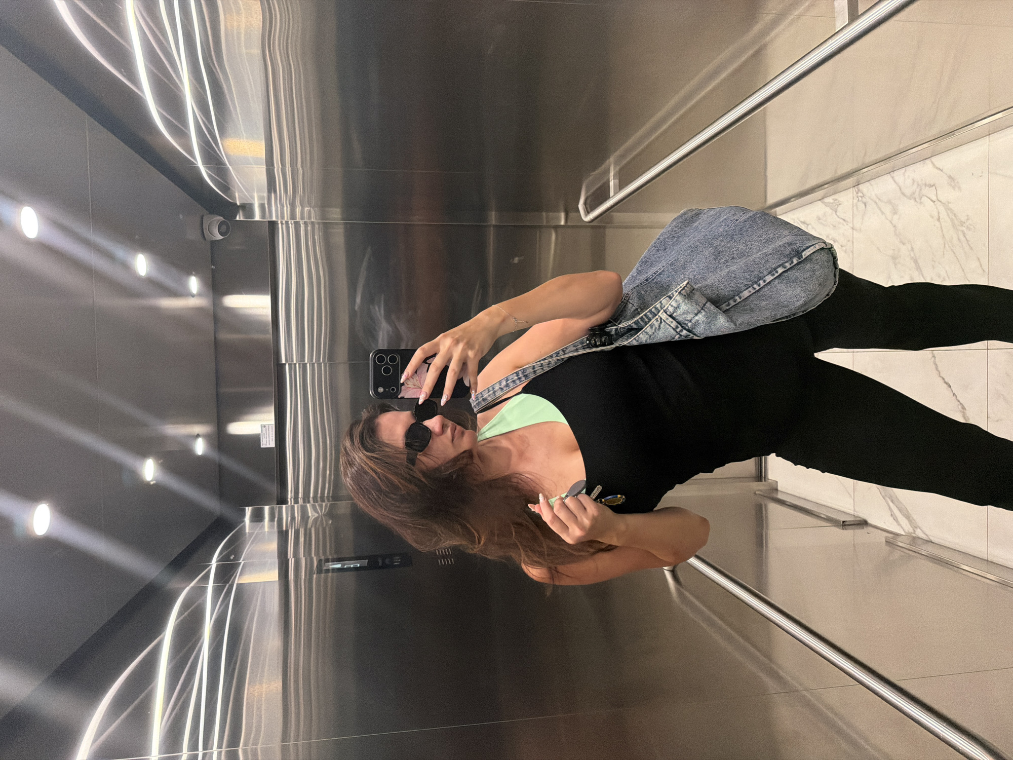 A woman is taking a mirror selfie in an elevator. She is wearing a black tank top over a mint green sports bra, black pants, and black sunglasses. She is holding her phone in her right hand and a set of keys in her left. A denim tote bag is slung over her left shoulder. The elevator has stainless steel walls and ceiling, with overhead lighting and a security camera visible. The floor is tiled with a white marble pattern. The scene is consistent with an elevator in a modern building, possibly in Belgrade, Serbia, given the context provided.