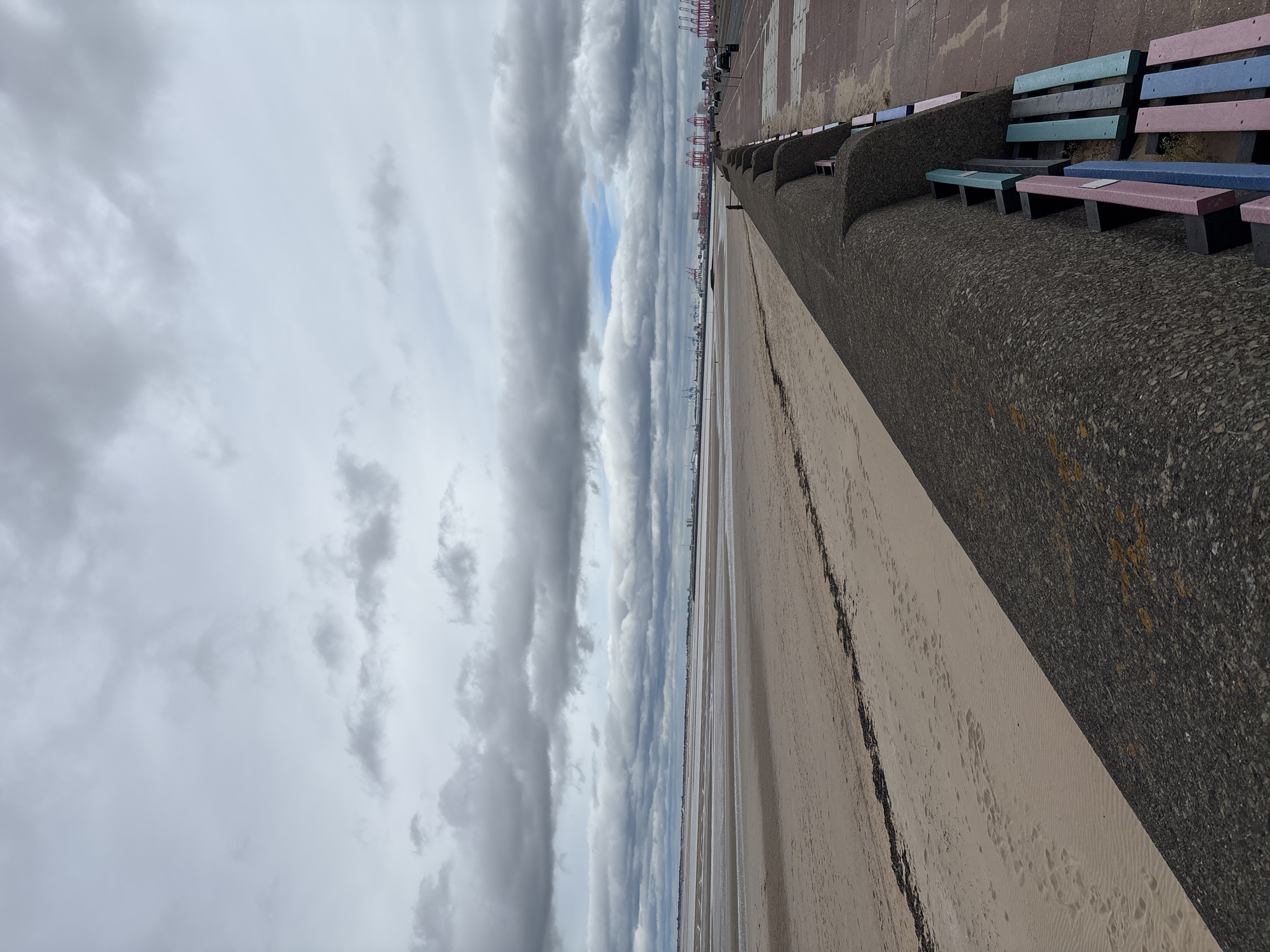 The image depicts a wide, sandy beach in Wallasey, United Kingdom, under a cloudy sky. In the foreground, a textured, dark grey concrete sea defense wall curves into the distance, with a series of brightly colored benches attached to its side. These benches are painted in various shades of pink, blue, and green. Beyond the wall is the expanse of the beach, showing ripples of sand and scattered dark seaweed. Further in the distance, across a vast expanse of water or sand at low tide, a city skyline is visible, characterized by numerous cranes and shipping containers, suggesting a busy port. A road runs alongside the sea defense wall, with a few cars visible. The overall scene is overcast and suggests a typical day at a coastal urban environment.