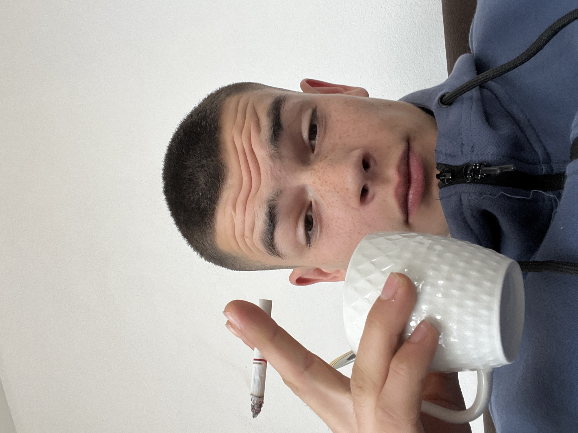 A young man with short, dark hair and freckles is holding a lit cigarette in one hand and a white, textured mug in the other. He is wearing a blue hooded sweatshirt. The background is a plain white wall. The location is not discernible from the image.