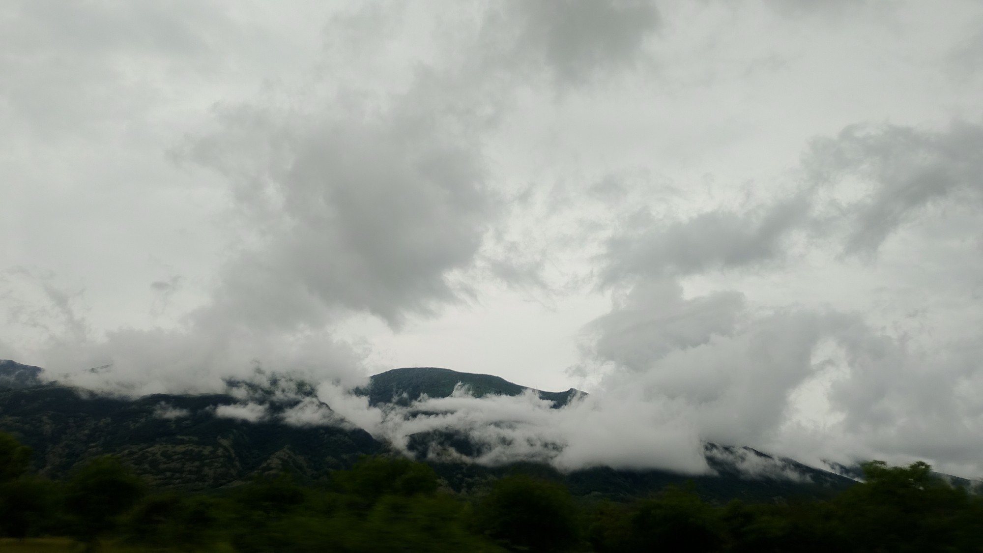 A low-angle, wide shot captures a natural landscape dominated by a cloudy, overcast sky and rolling, tree-covered mountains. The clouds are dense and dark, creating a somewhat muted and dramatic atmosphere. Wisps of fog and low-lying clouds partially obscure the peaks and slopes of the mountains, suggesting a humid or misty environment. The lower portion of the image shows the tops of dark green trees, hinting at a dense forest or foliage. The scene appears to be a mountainous region with substantial cloud cover. There are no people or man-made structures visible in the frame, indicating a natural and undeveloped setting.