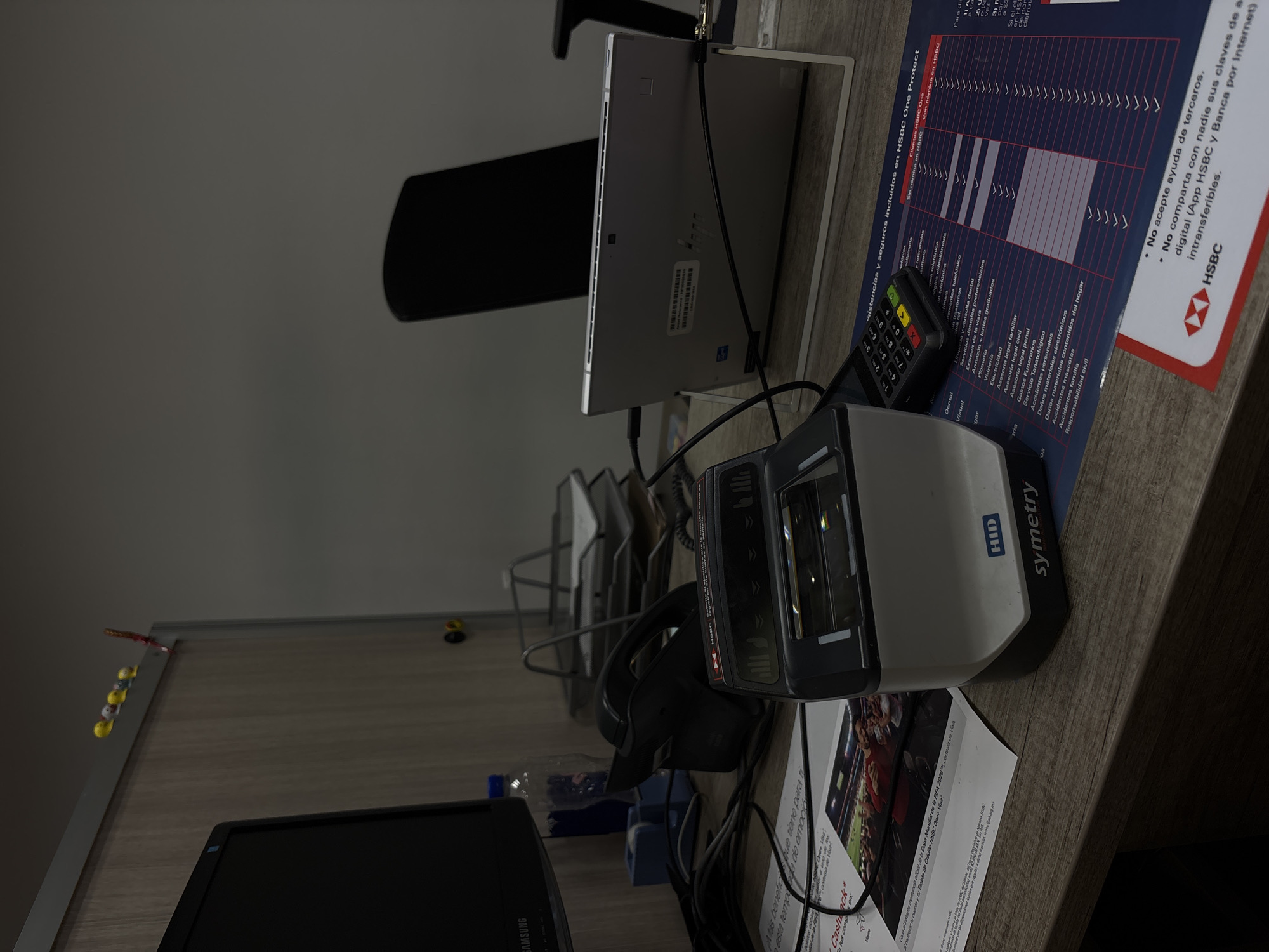 The image depicts a desk setting, likely in an office environment, with various objects placed on its surface. A black Samsung monitor is positioned at the bottom left corner. Adjacent to it is a clear plastic bottle with blue cap. A white and grey barcode scanner is present, partially obscuring a document with the word "Cashback" printed on it, along with images of a soccer stadium and the FIFA World Cup logo. A black credit card terminal is nearby. A silver tablet or laptop, standing in a metal frame, is visible in the background, along with a black office chair. An HSBC document showing "HSBC One Protect" insurance benefits and an offer to get official merchandise from FIFA 2026 is on the upper right corner. A pencil holder with pencils and erasers on the corner of the table. The environment appears to be an office, possibly in Mexico City, given the presence of the HSBC document with Spanish text.